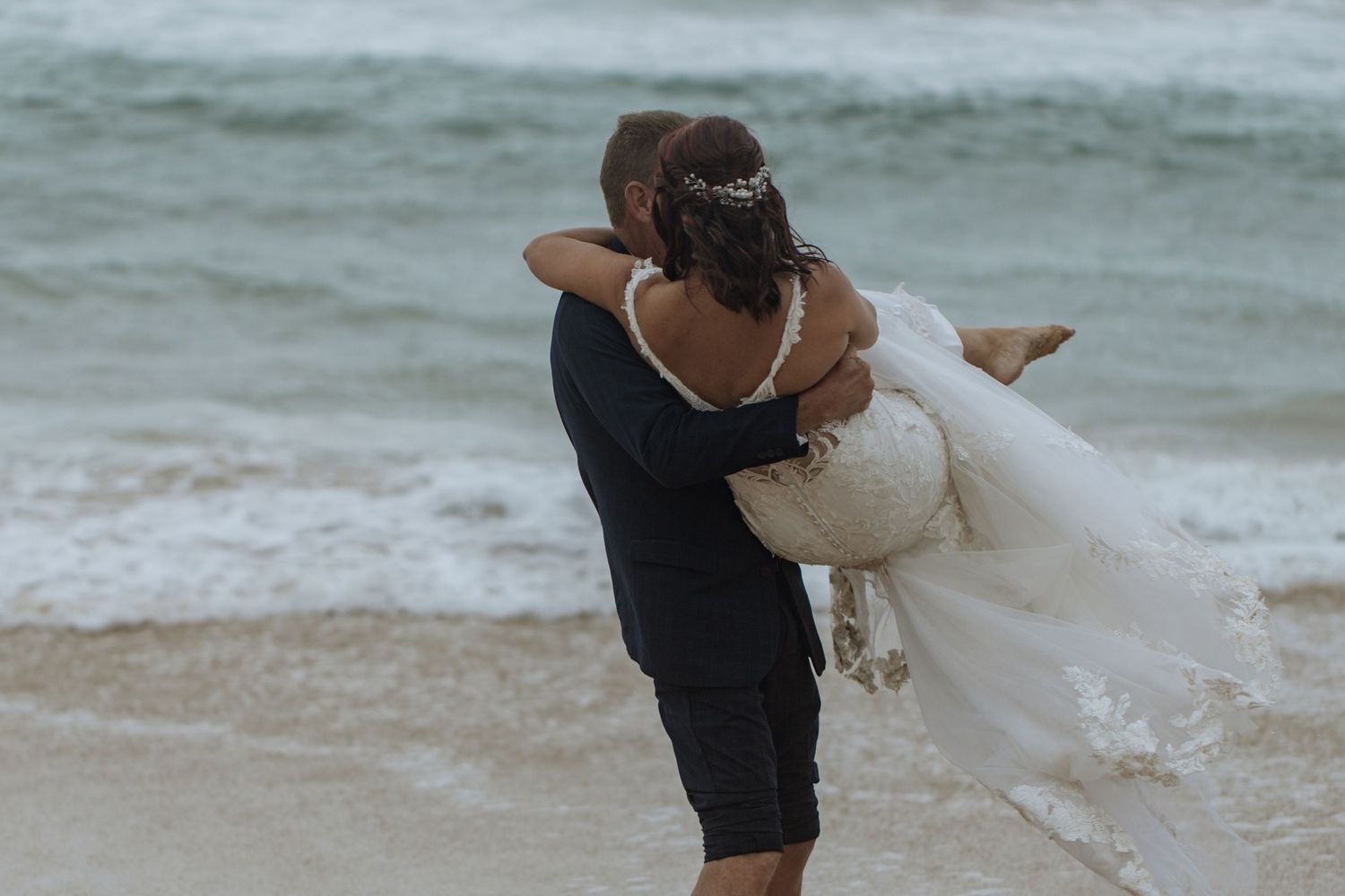 Couple embraces in a romantic moment at the beach with waves crashing behind them.