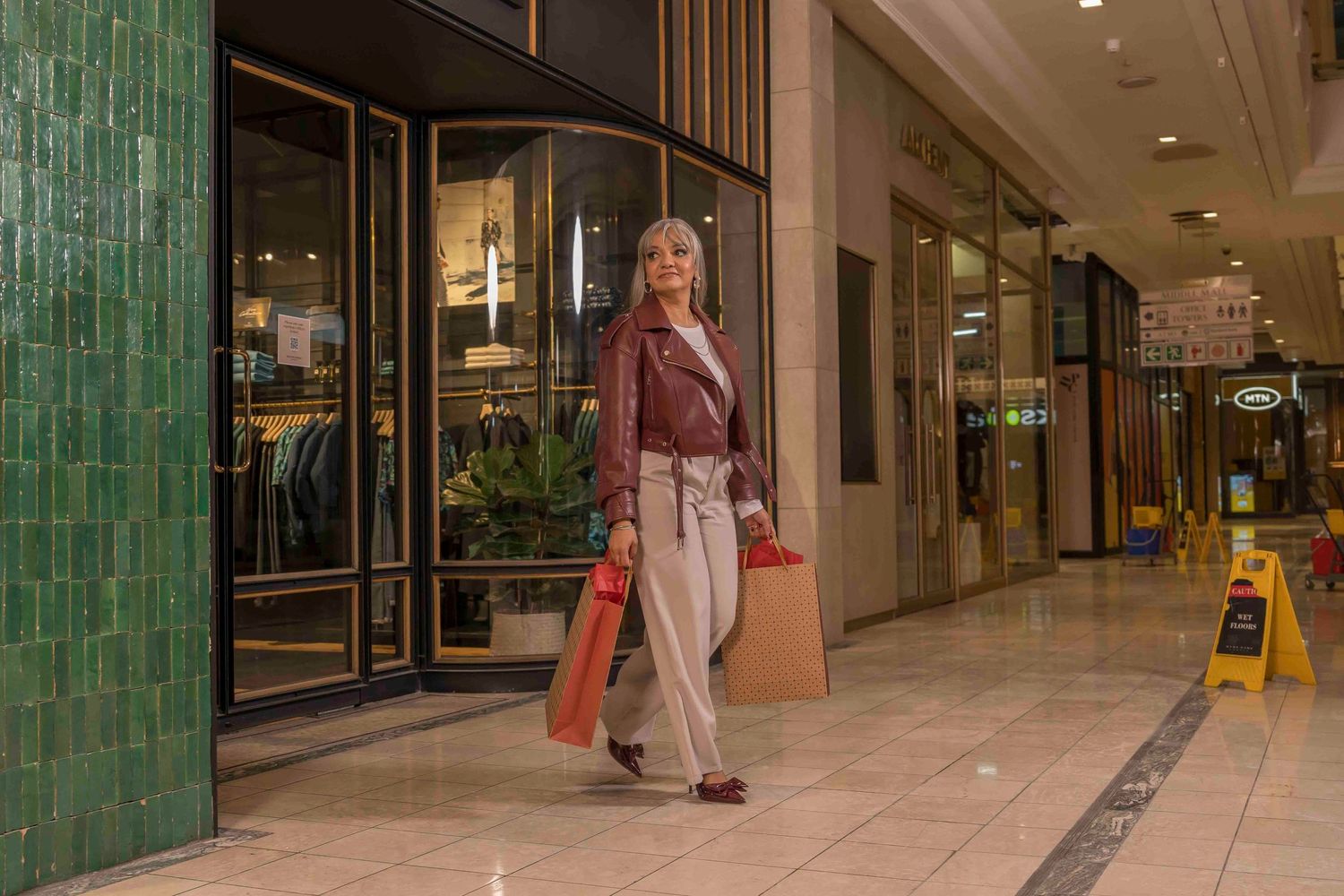 Shopper with bags walks past retail stores in a mall corridor with green tiled pillars and yellow caution sign.