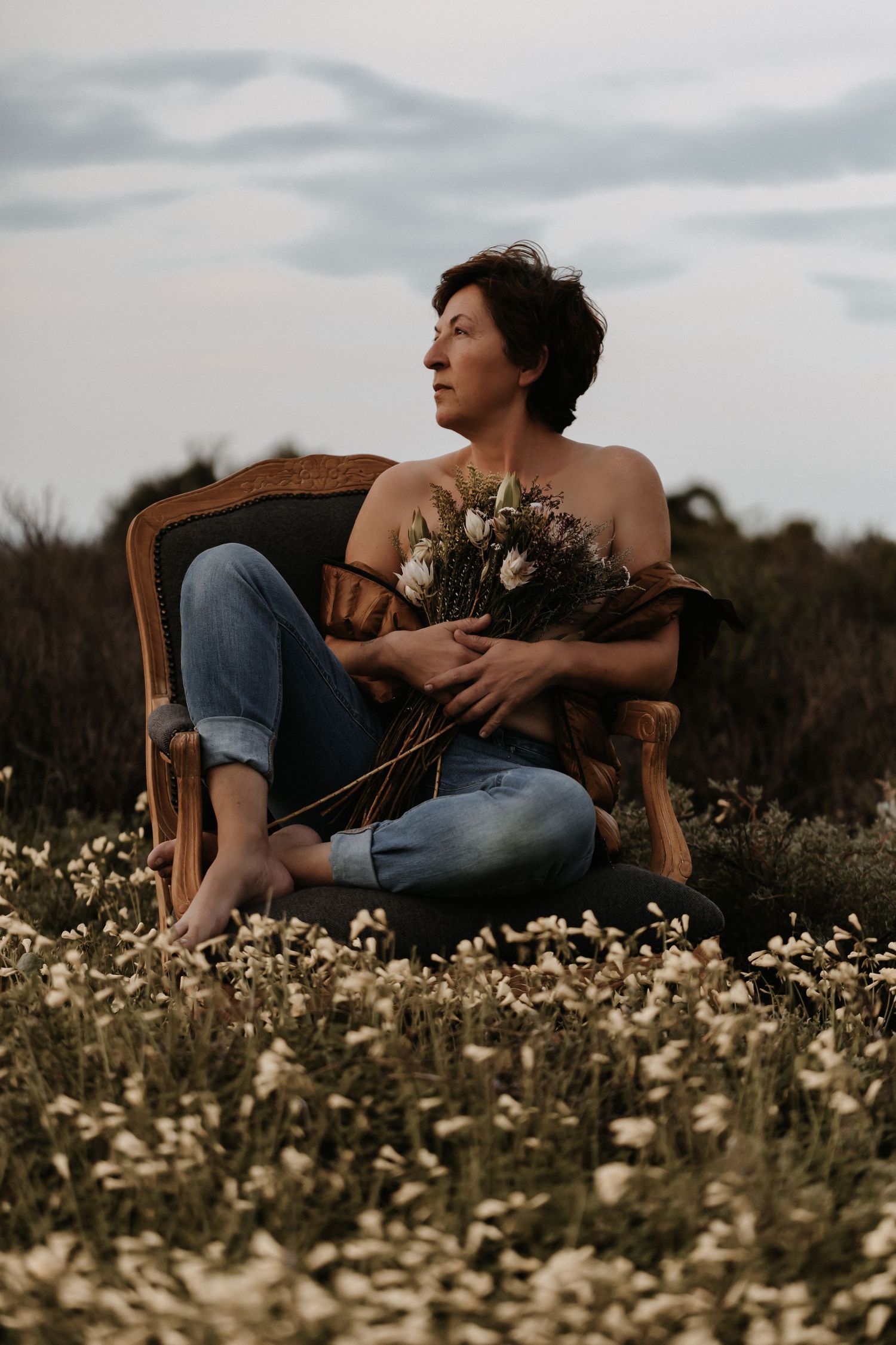 A person sits barefoot in a vintage chair among wildflowers holding a bouquet during sunset.