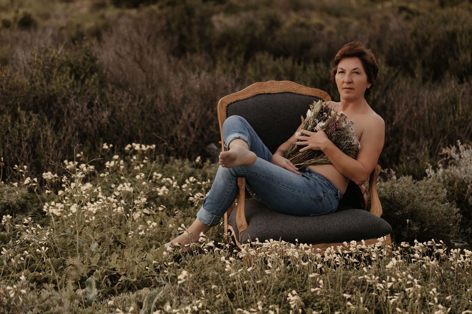 A relaxed portrait in a vintage armchair surrounded by wildflowers in a rustic outdoor setting during golden hour.