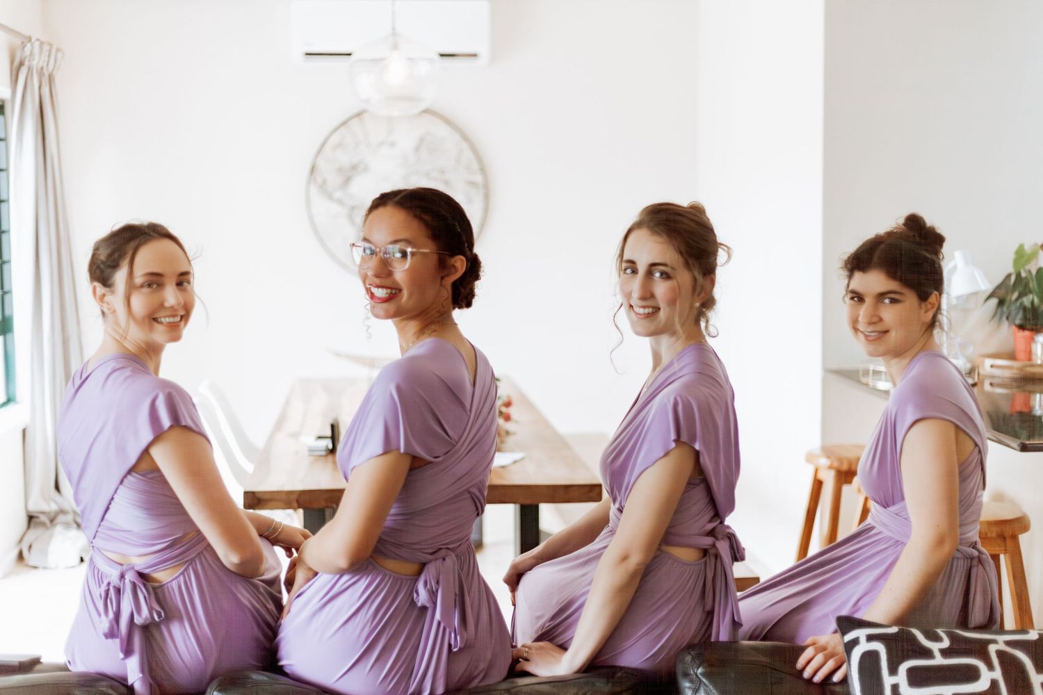 Four bridesmaids in matching lilac dresses sit at a table looking over their shoulders at the camera.