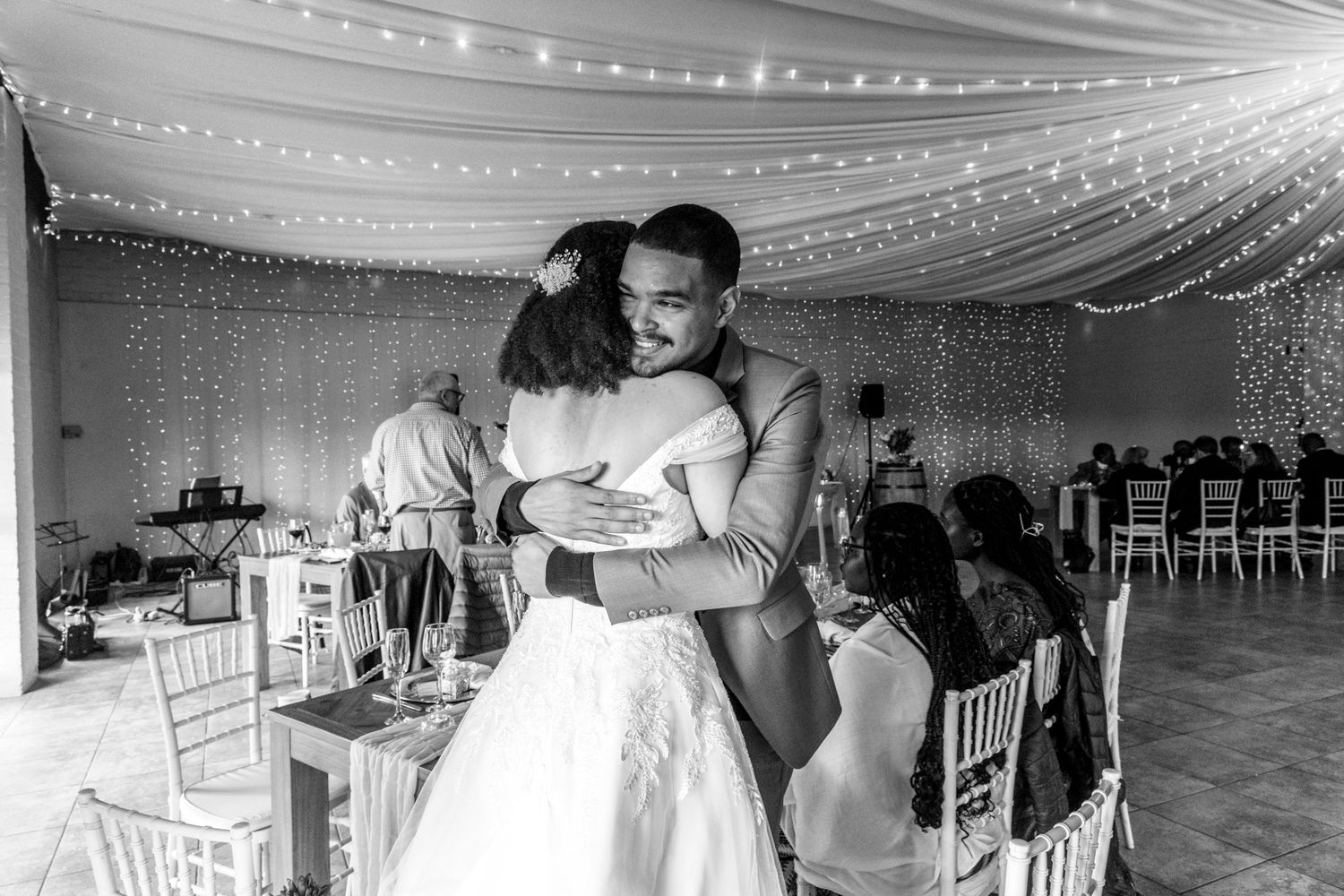 A tender embrace between two people dancing at an elegant wedding reception under twinkling string lights.