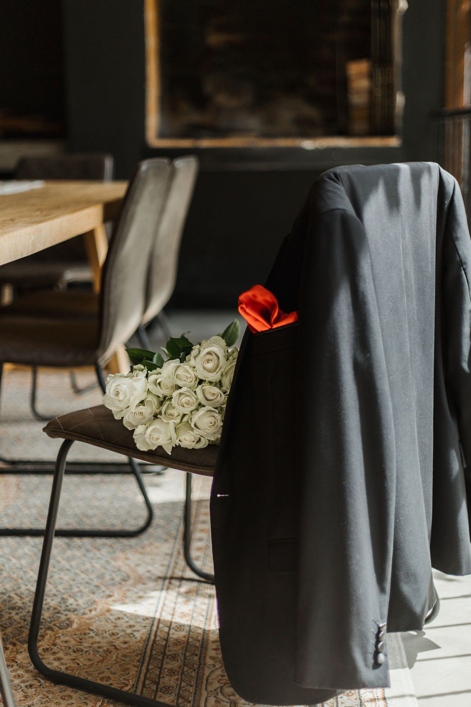 Dark suit jacket with red pocket square draped over chair beside white roses bouquet in elegant interior setting.