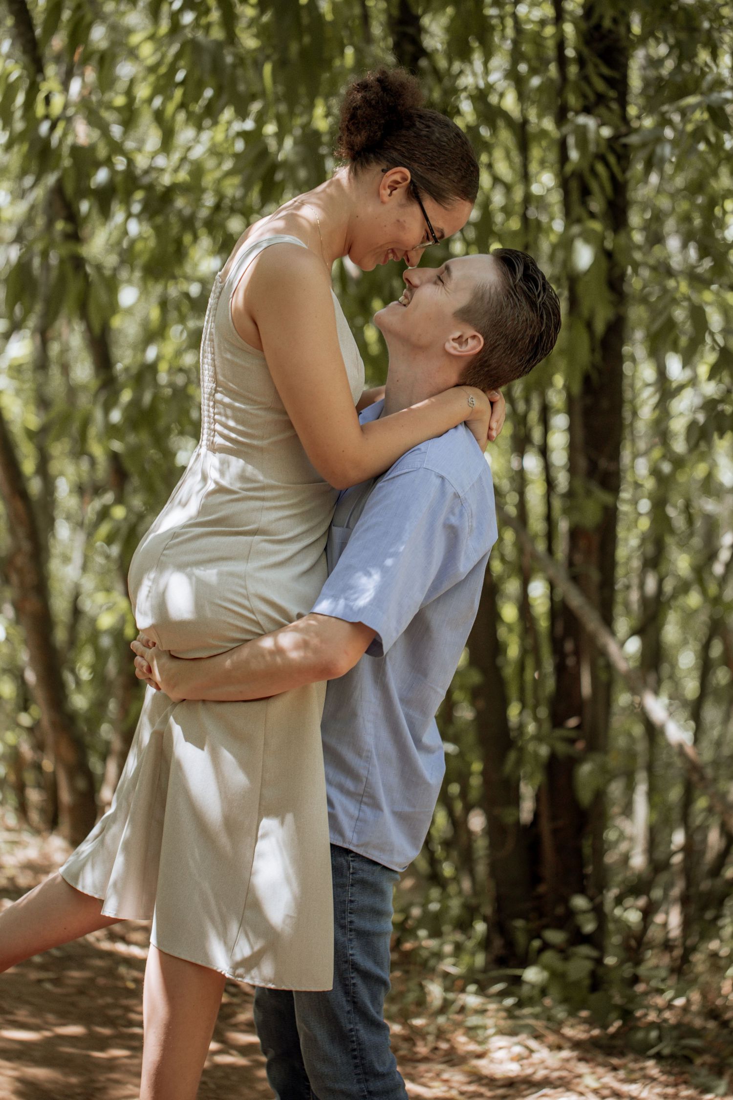 A romantic couple embraces in a sunlit forest setting, with the woman in a light dress being lifted by her partner.