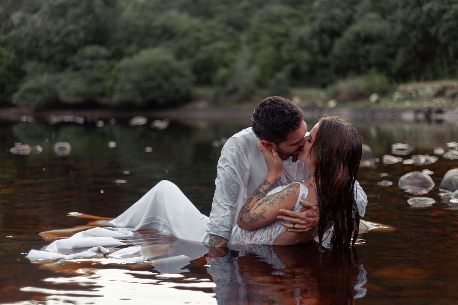 A romantic couple embraces while standing in a shallow river surrounded by rocks and trees.