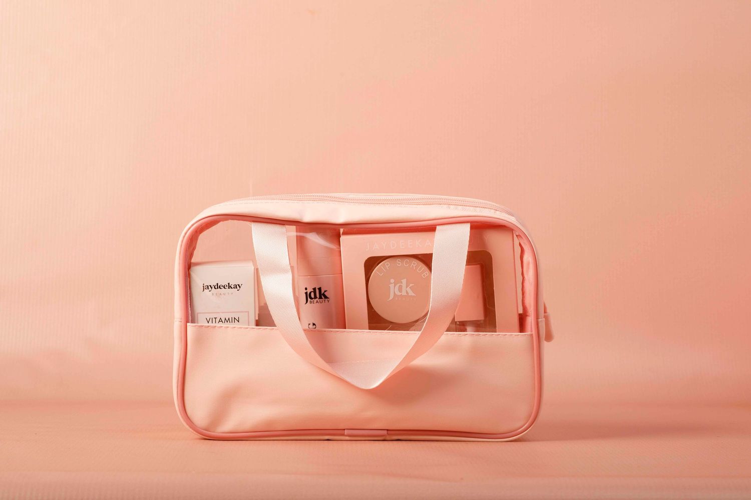 Pink tinted cosmetic bag with clear panels and zipper closure displayed on a peachy pink background.