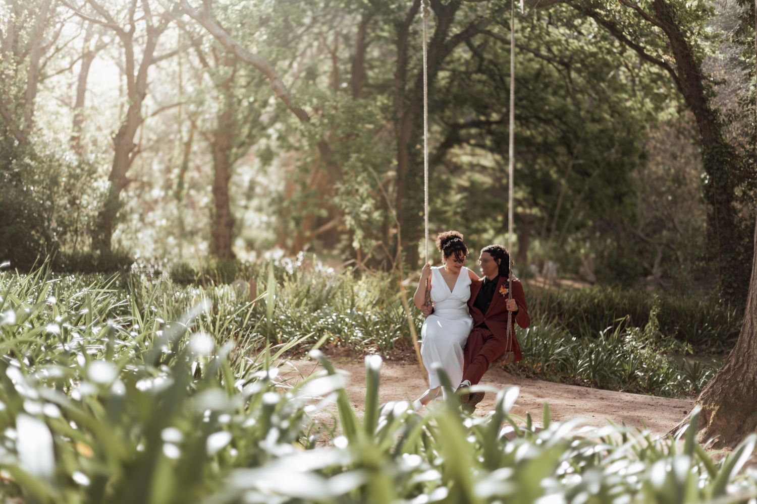 Two people hold hands and walk down a romantic tree-lined path covered in white flower petals during a sunny day.