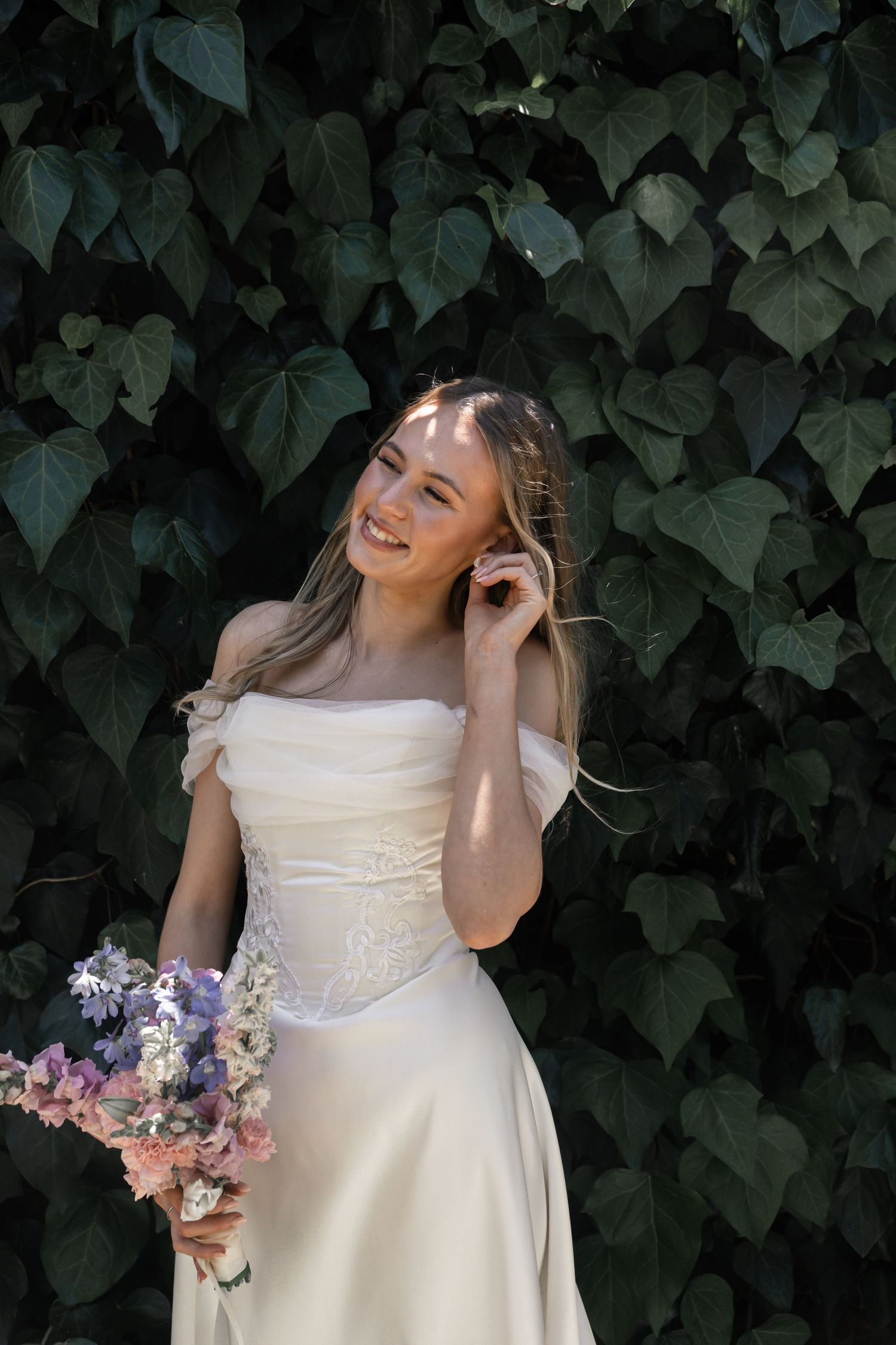 A person in a cream-colored dress poses joyfully against a backdrop of dark green ivy leaves, holding a pastel bouquet.