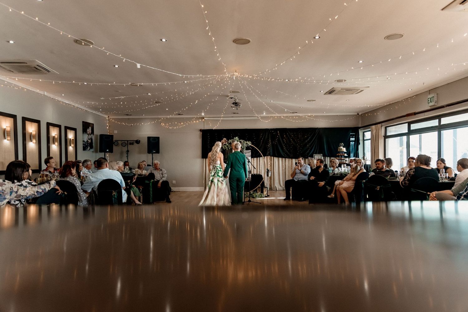 A wedding ceremony takes place in an elegant reception hall with string lights and guests seated at round tables.