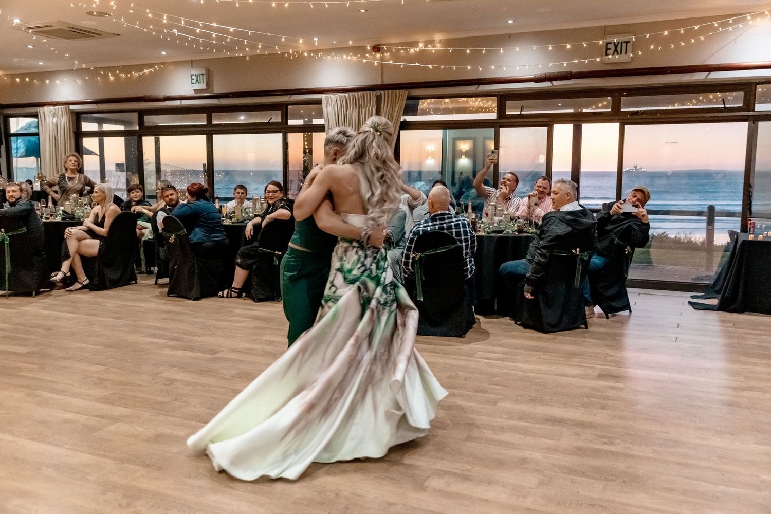 A couple dancing at a wedding reception in a venue with large windows overlooking the ocean at sunset.