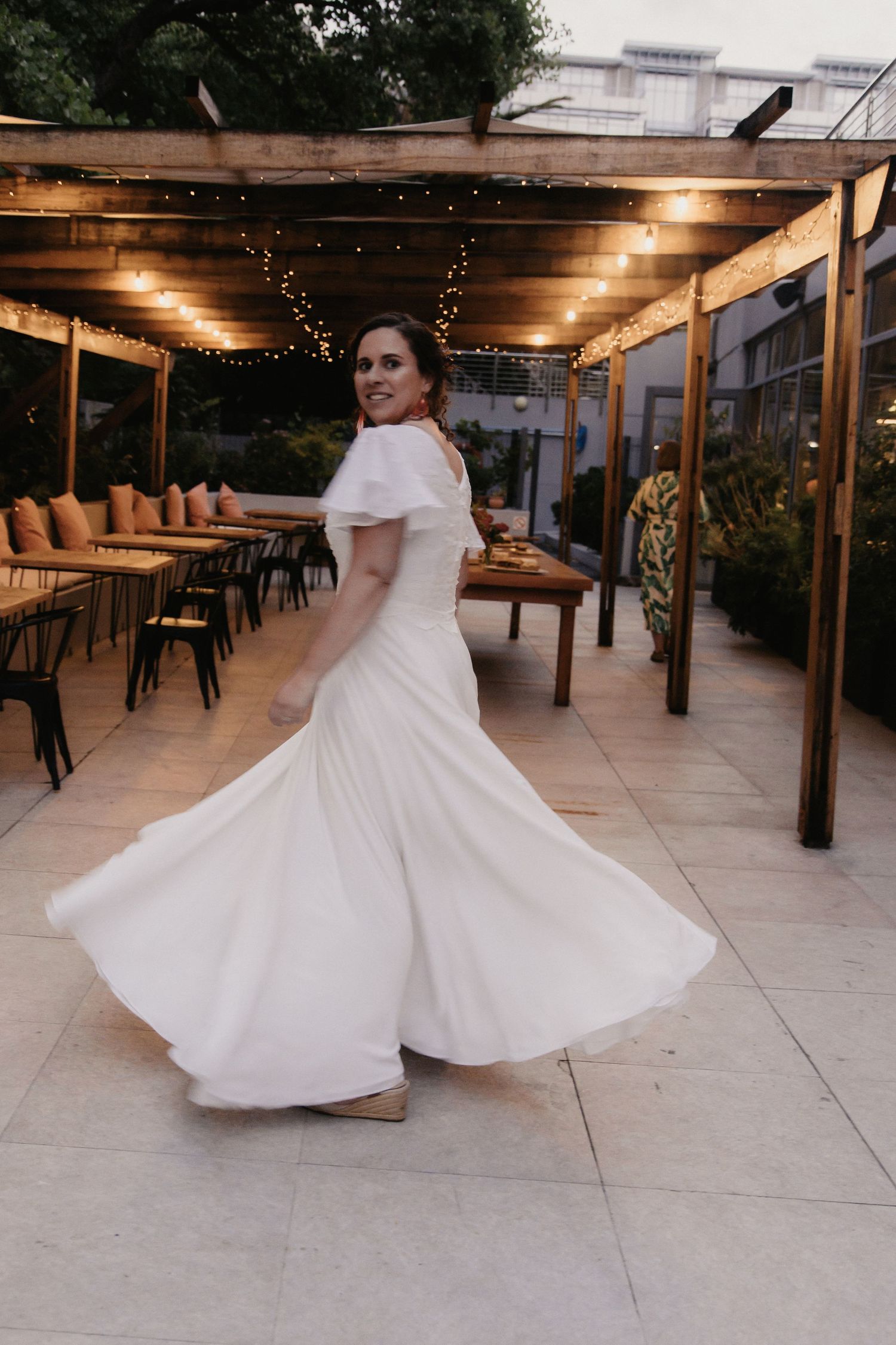 A bride in a flowing white dress twirls and spins on an outdoor patio under string lights at dusk.