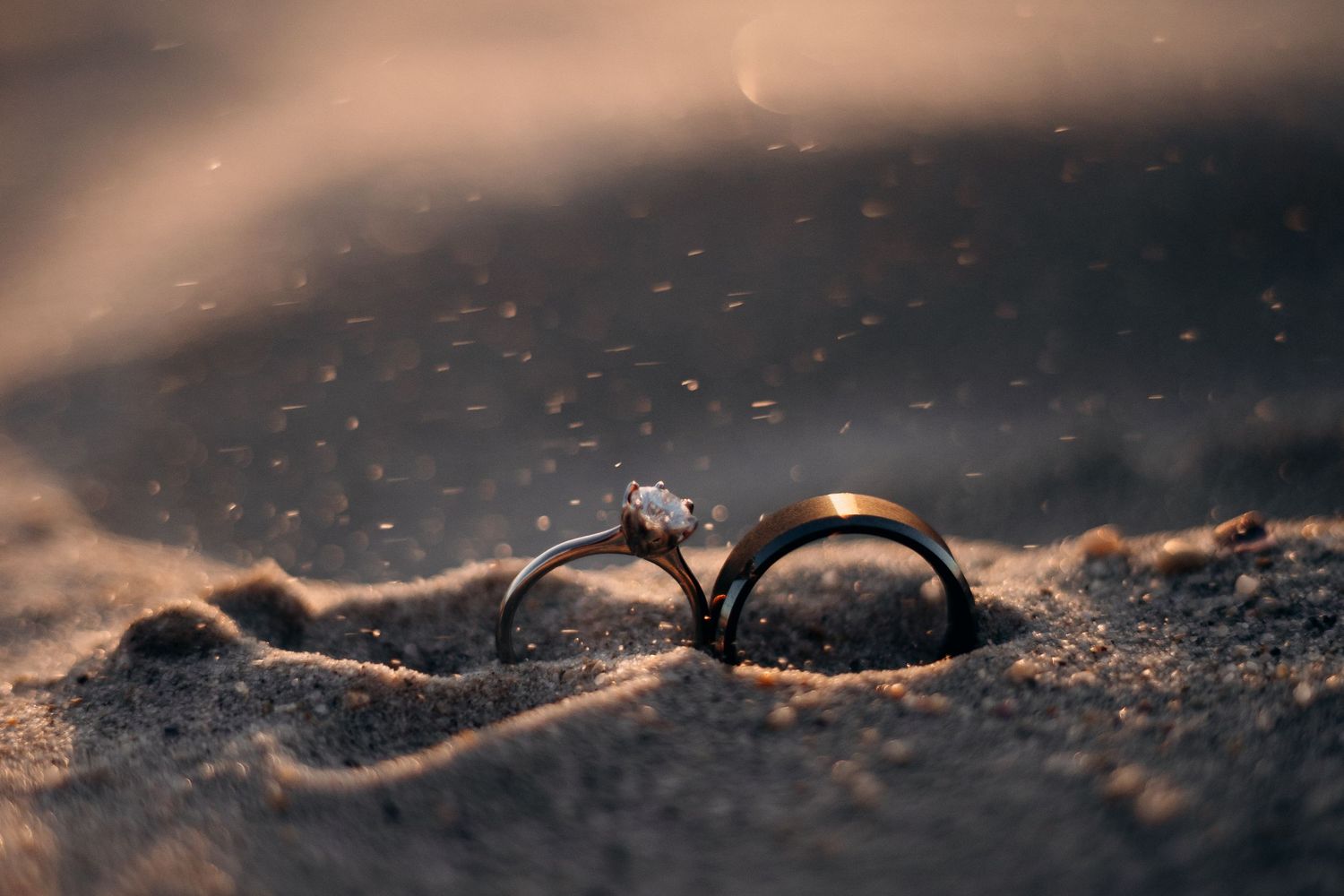 Two wedding rings, one with a diamond, resting in golden sand with bokeh light particles in the background.