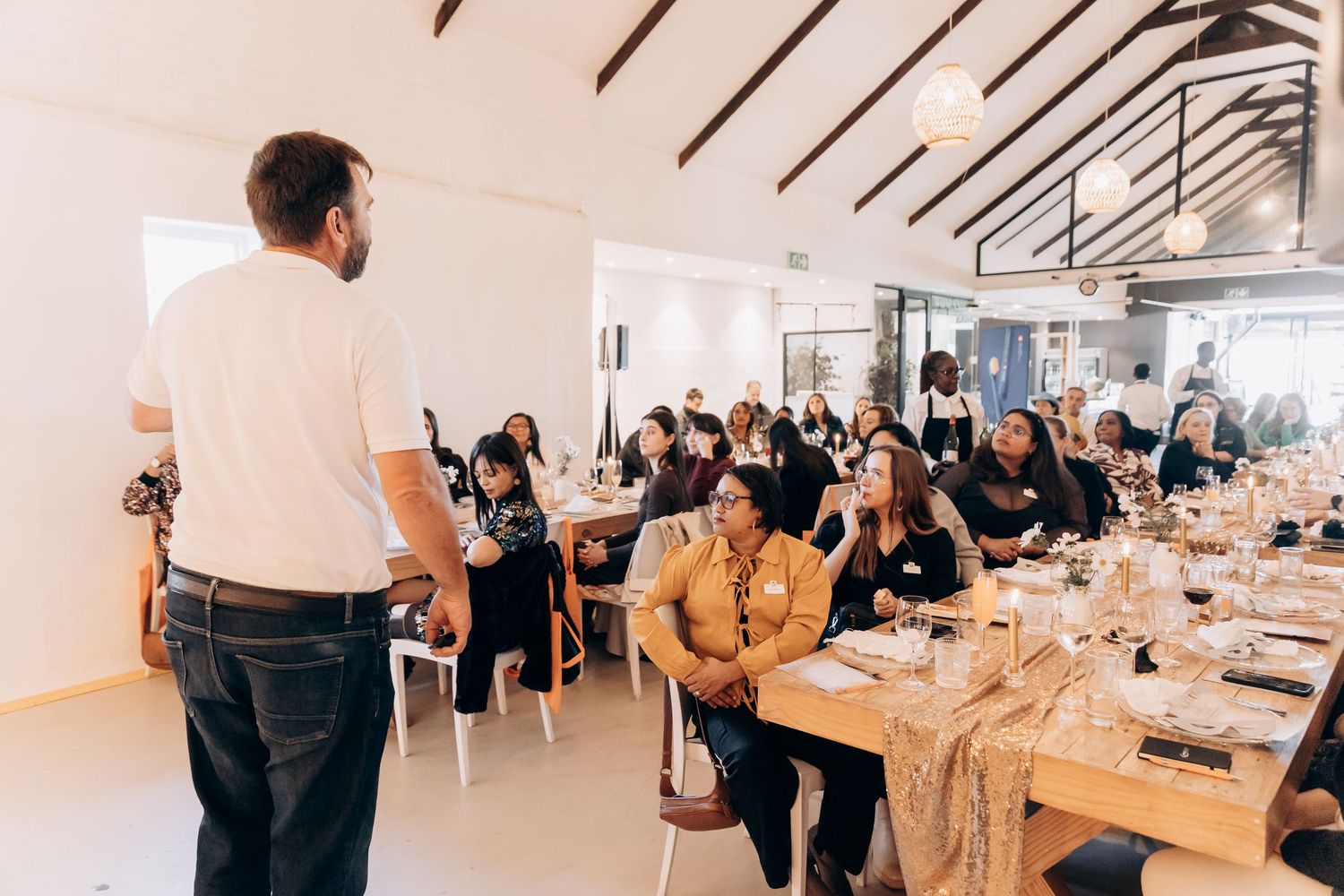 A man presents to a diverse audience seated at elegantly set tables in a bright, modern event venue.
