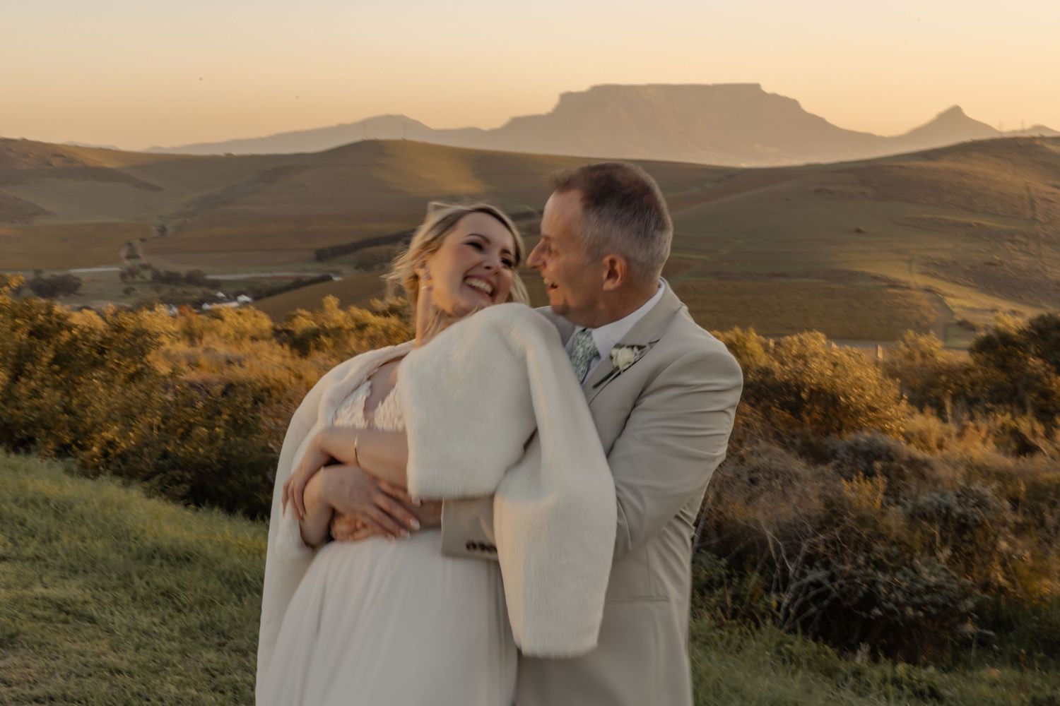 A couple embraces during golden hour sunset with scenic mountain landscape in the background.