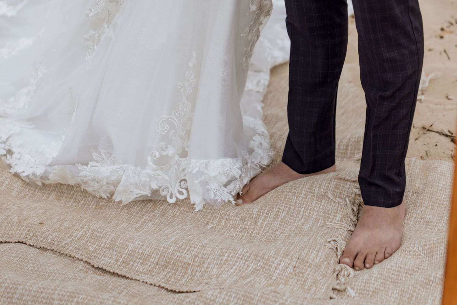 A closeup of bare feet standing next to the hem of a white lace wedding dress on a burlap rug.