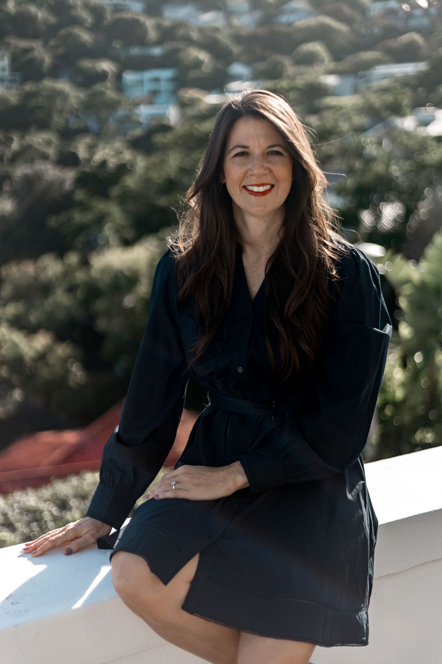 A young woman in a navy blue wrap dress sits casually on a balcony rail with sunny mountains in the background.
