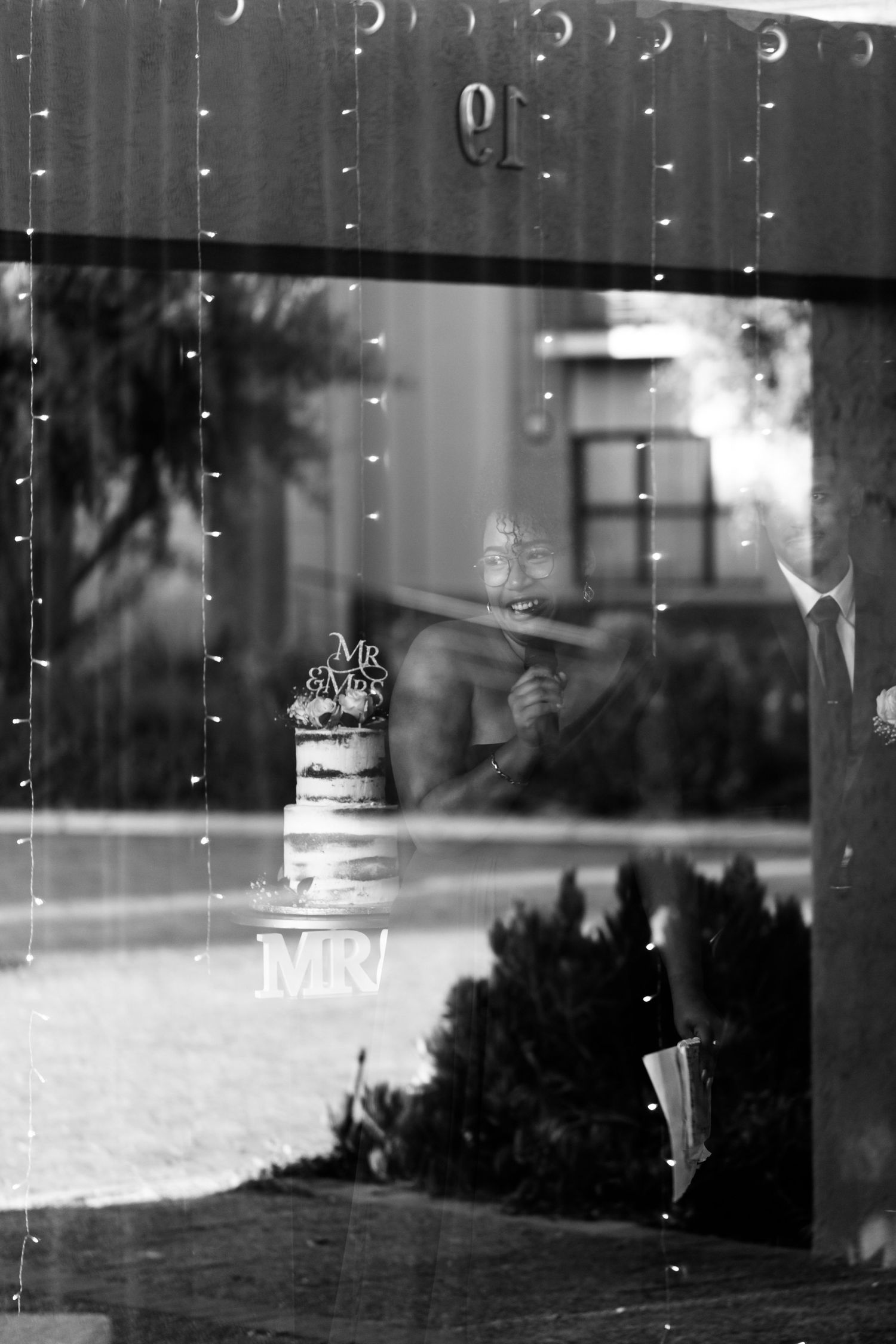 A black and white reflection through a window shows Mr & Mrs signage and a wedding cake in ethereal lighting.