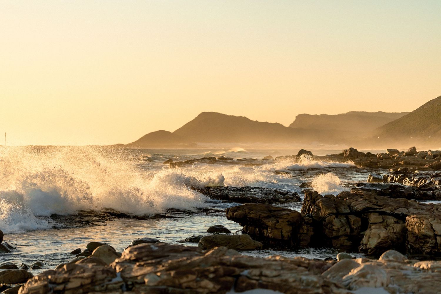 Ocean waves crash against rocky coastline at sunset with mountains silhouetted in the golden light.