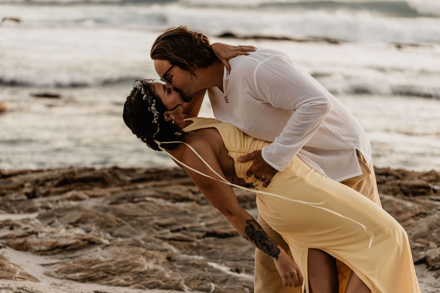 A romantic couple embraces and kisses on a rocky beach at sunset wearing flowy white and beige outfits.
