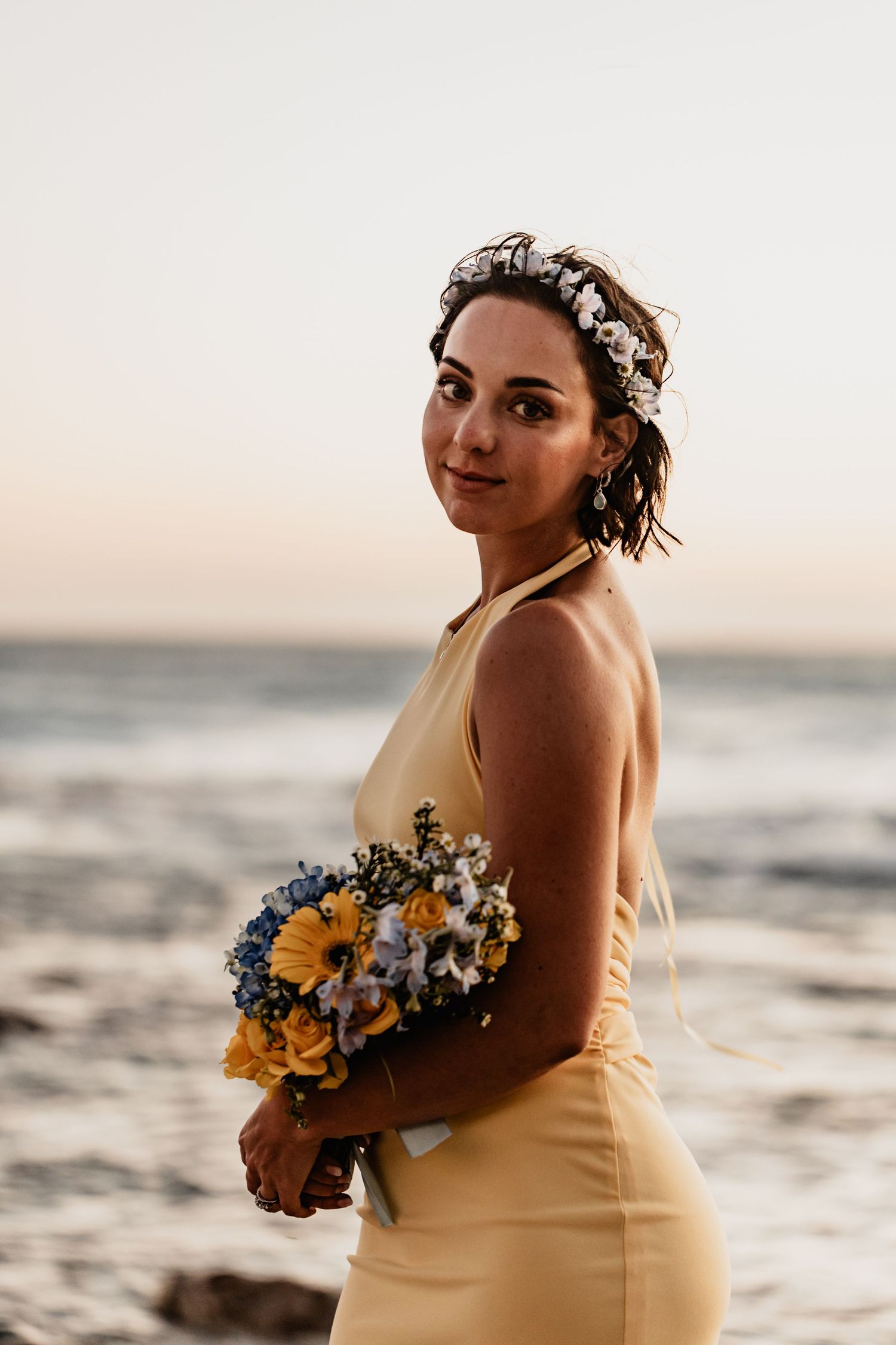 A woman in a yellow dress holds a bouquet of sunflowers and blue flowers against a sunset beach backdrop.