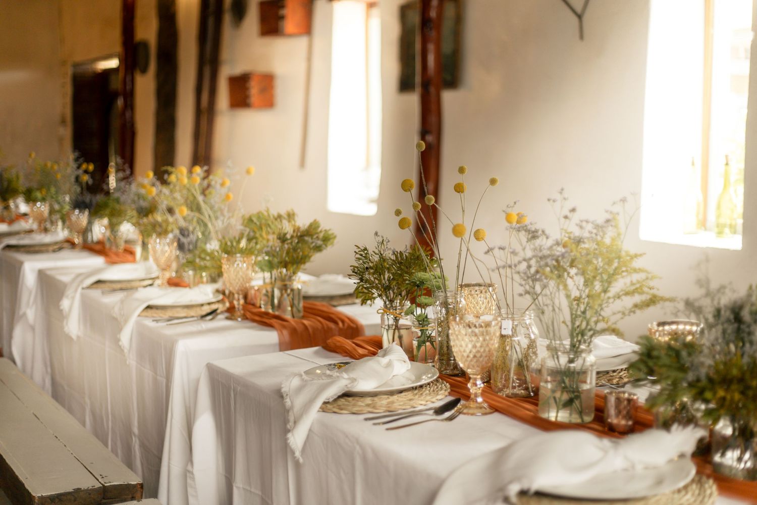 Elegant rustic wedding reception table with white linen, terracotta runners, wildflower arrangements, and gold accents in a barn venue.