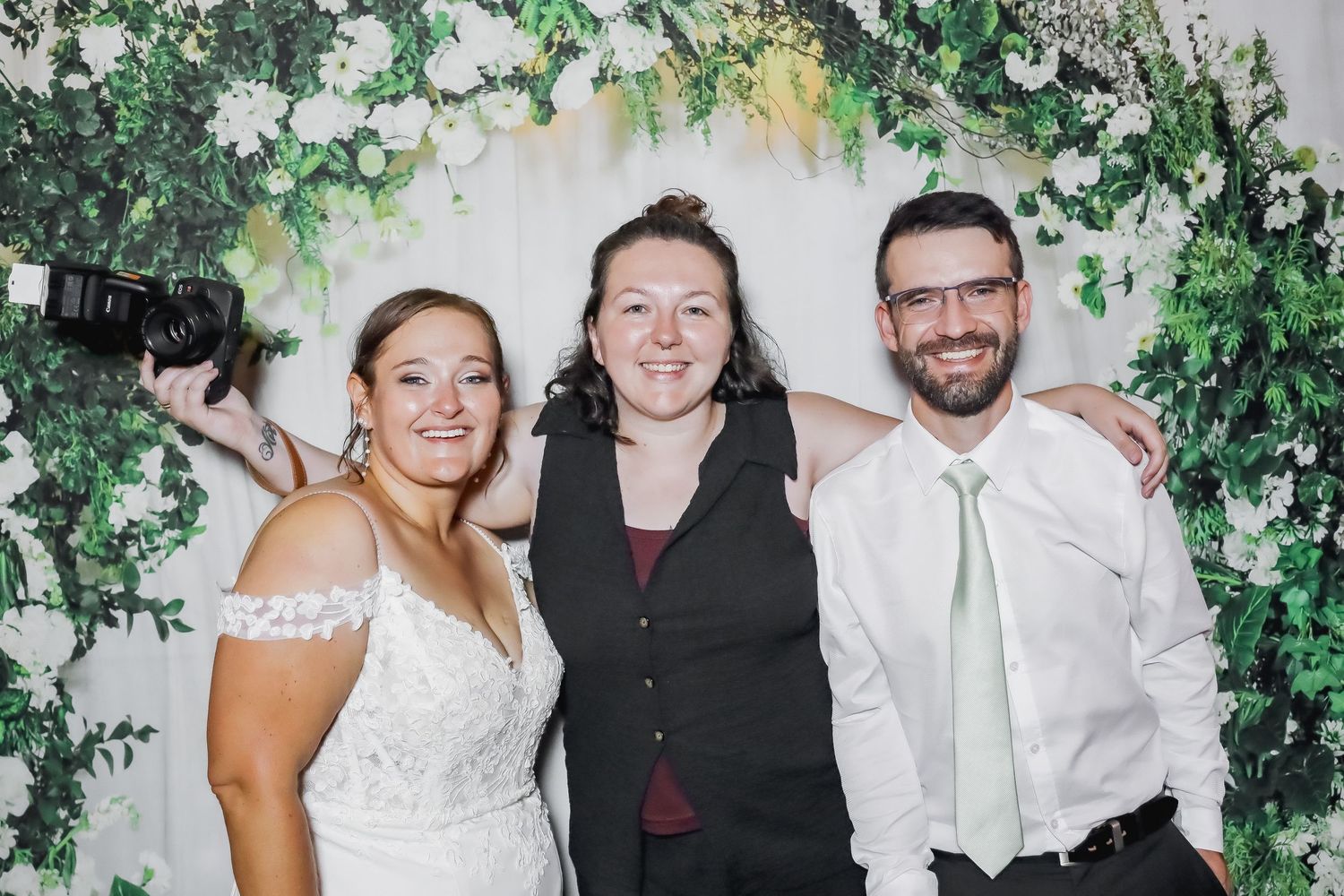 Bride in white gown and groom in white shirt with green tie pose with wedding photographer under floral arch.