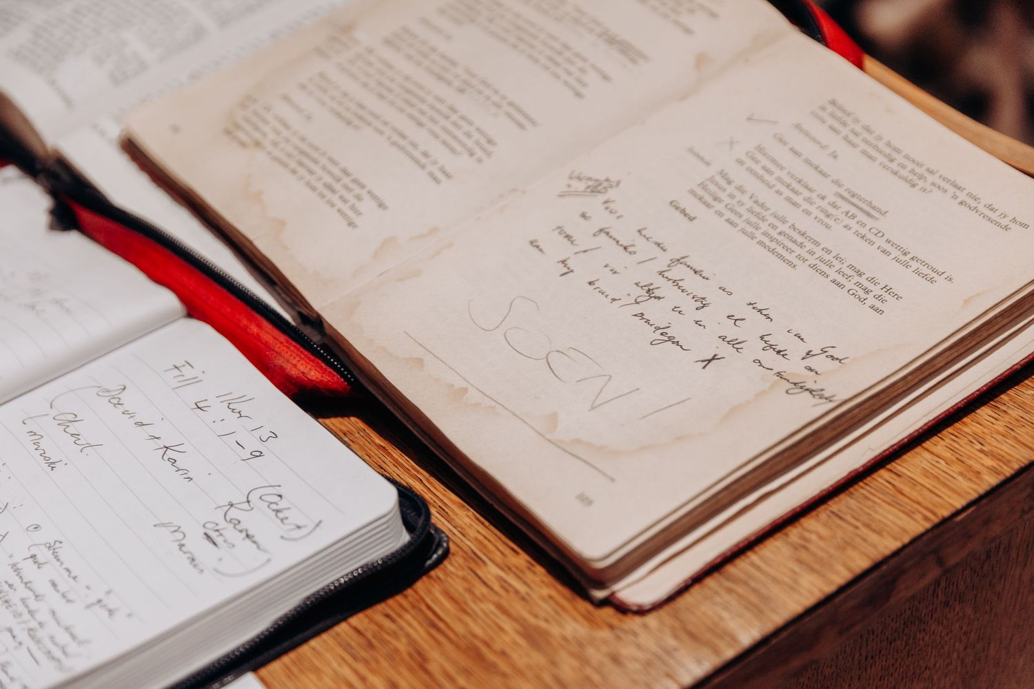 Open journals with handwritten notes and printed text rest on a wooden surface with red ribbon bookmarks.