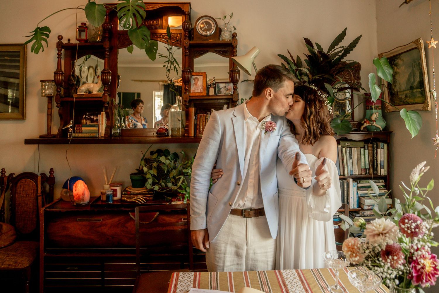 Couple sharing a romantic kiss in a boho-styled room with lush plants, wooden shelves, and floral arrangements on their wedding day.