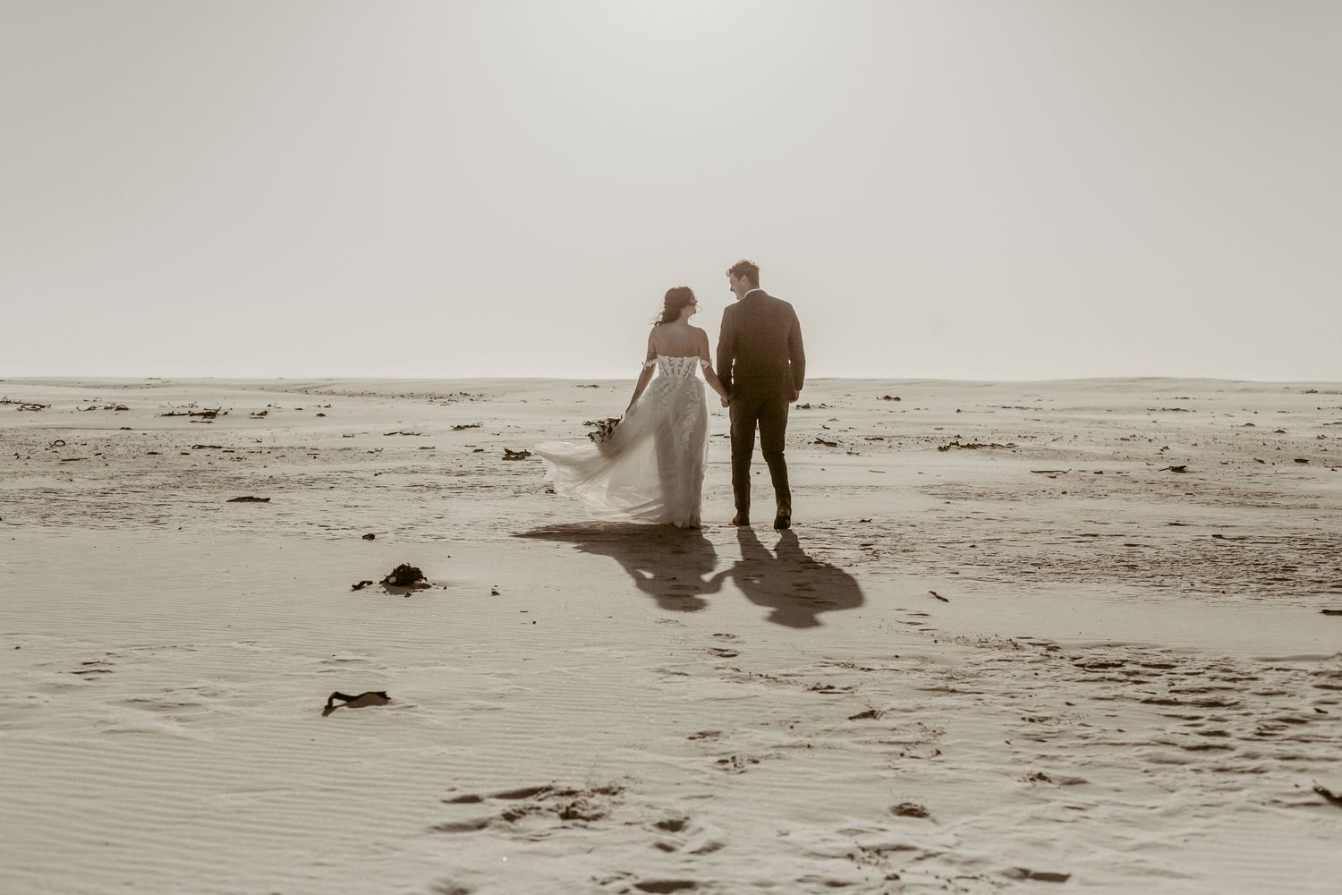 Newlywed couple standing together on sandy beach in wedding attire facing the ocean in soft sepia tones.