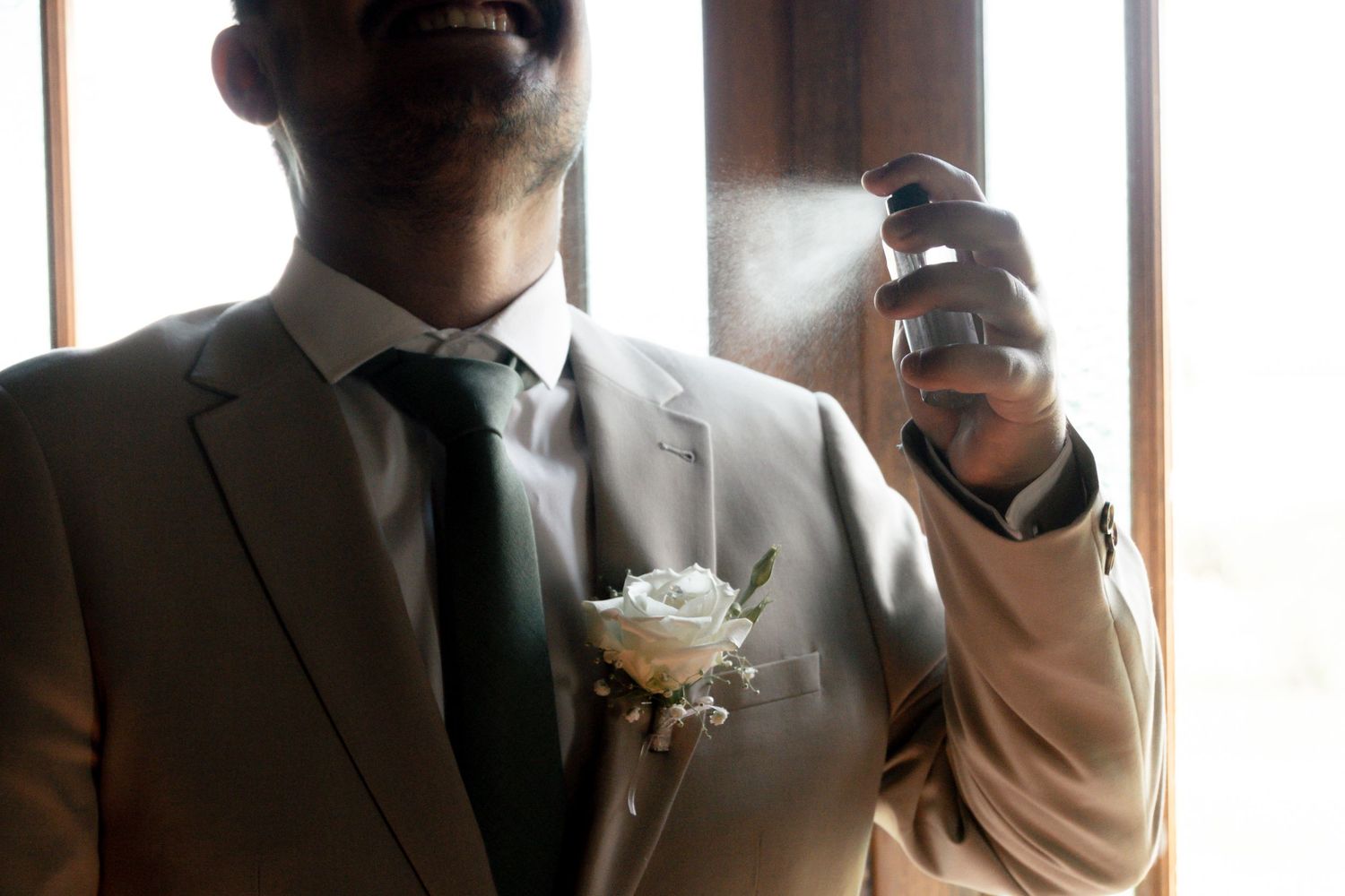 Groom in gray suit with white boutonniere spraying cologne before wedding ceremony.