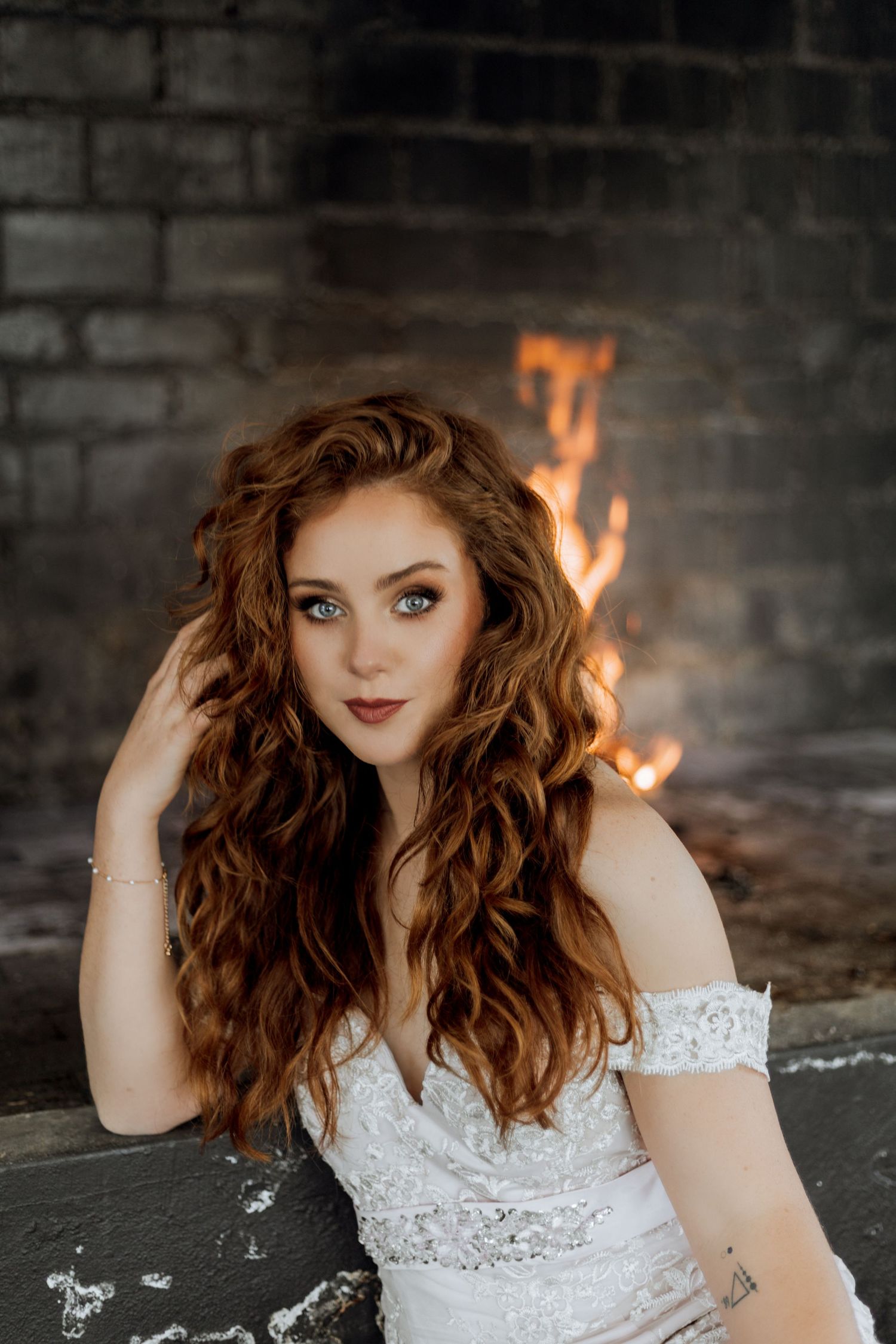 Fashion portrait of a woman with curly red hair in a white lace dress against a dark brick wall with fire elements.