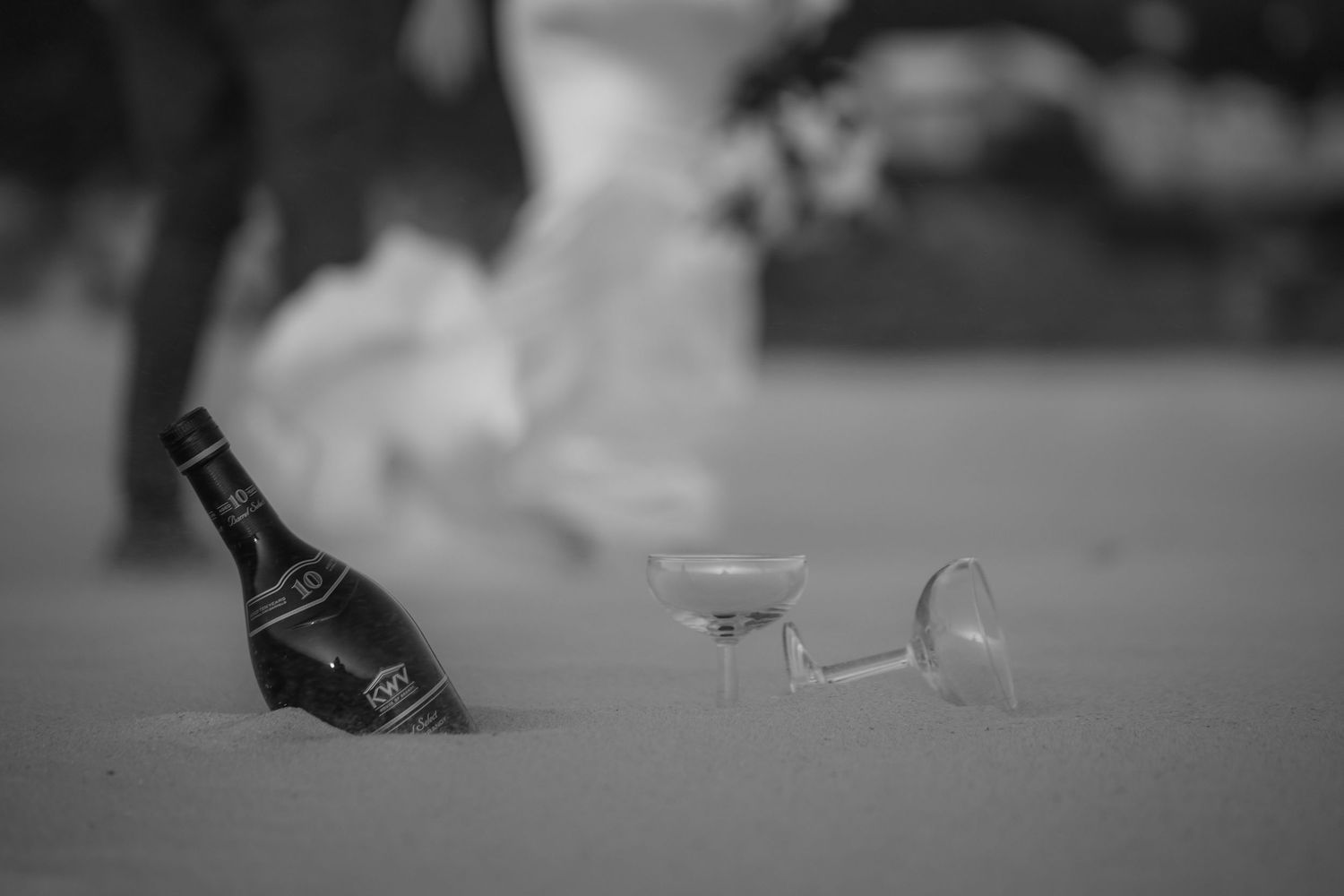 Black and white photo of a wine bottle and two glasses lying in sand at a beach wedding celebration.