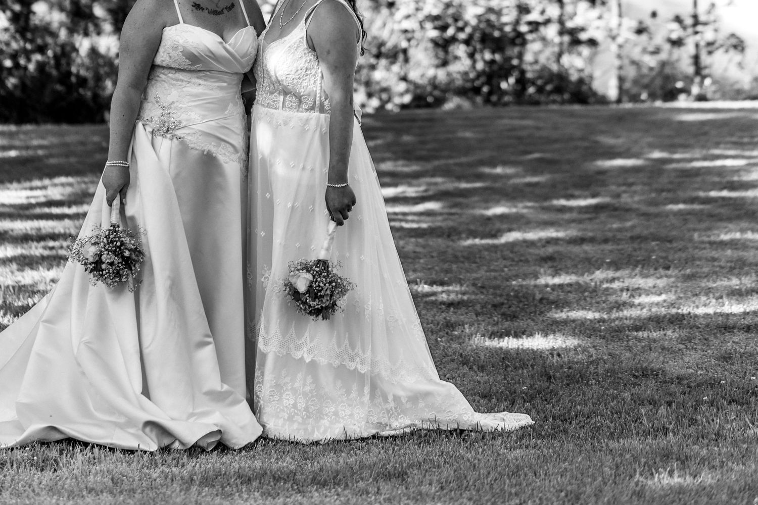 Black and white close-up of two brides in white wedding dresses holding bouquets together on grass.
