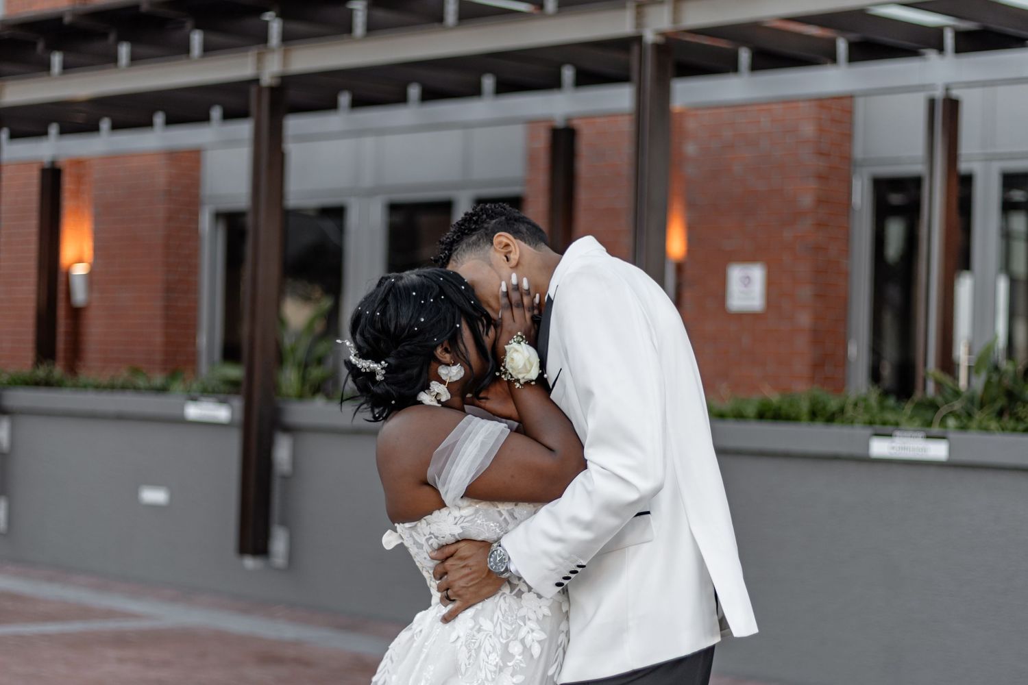A newlywed couple shares a romantic kiss during their wedding photoshoot outside a brick building.