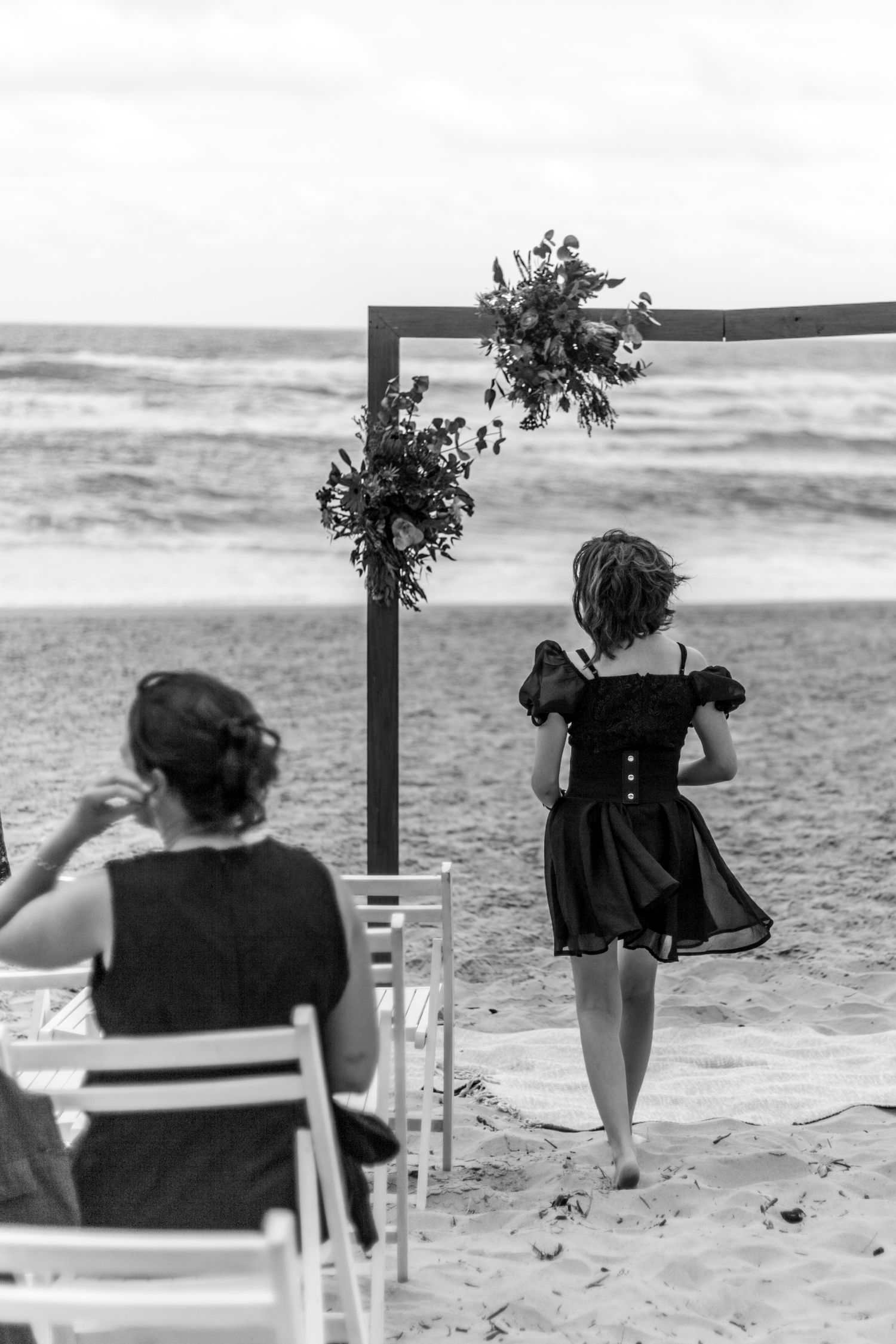 Black and white beach wedding scene with floral arch and chairs on the sand as waves roll in along the shoreline.