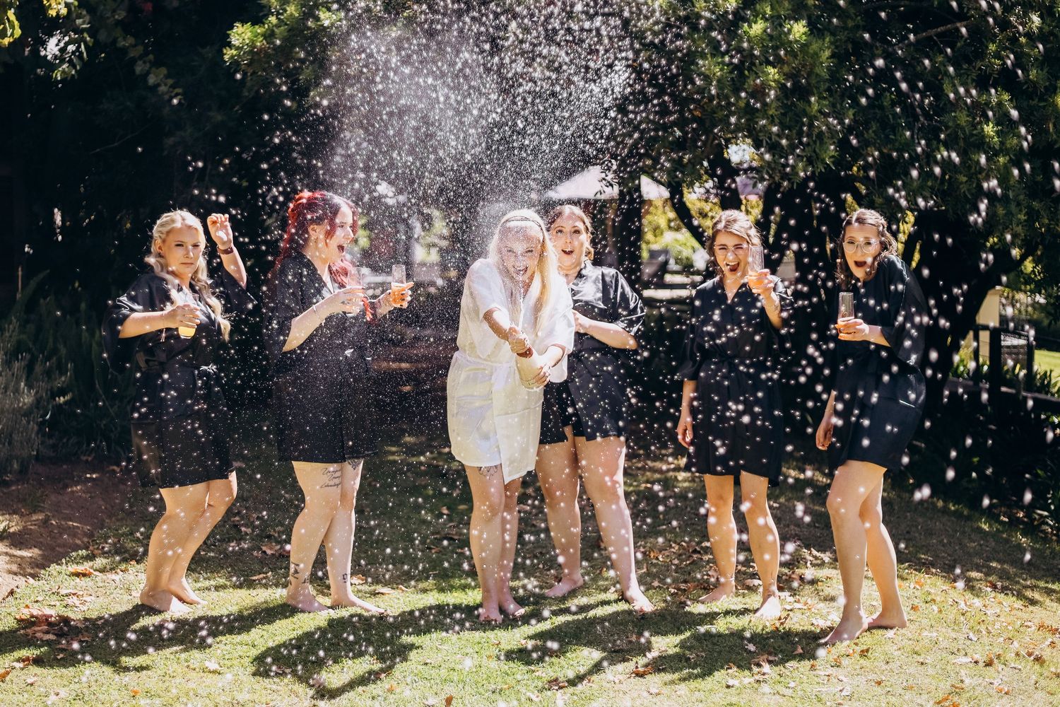 Bride and bridesmaids in navy robes celebrate with champagne spray outdoors on a sunny wedding day.
