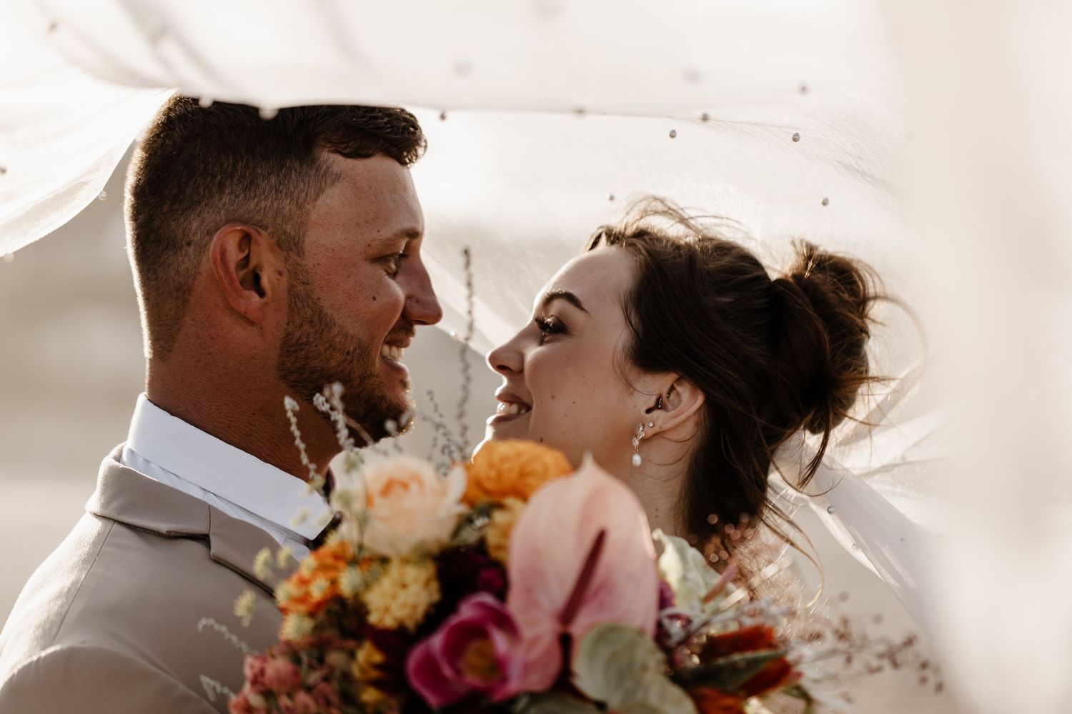 A joyful bride and groom share an intimate moment under a veil, with a colorful bridal bouquet in hand.