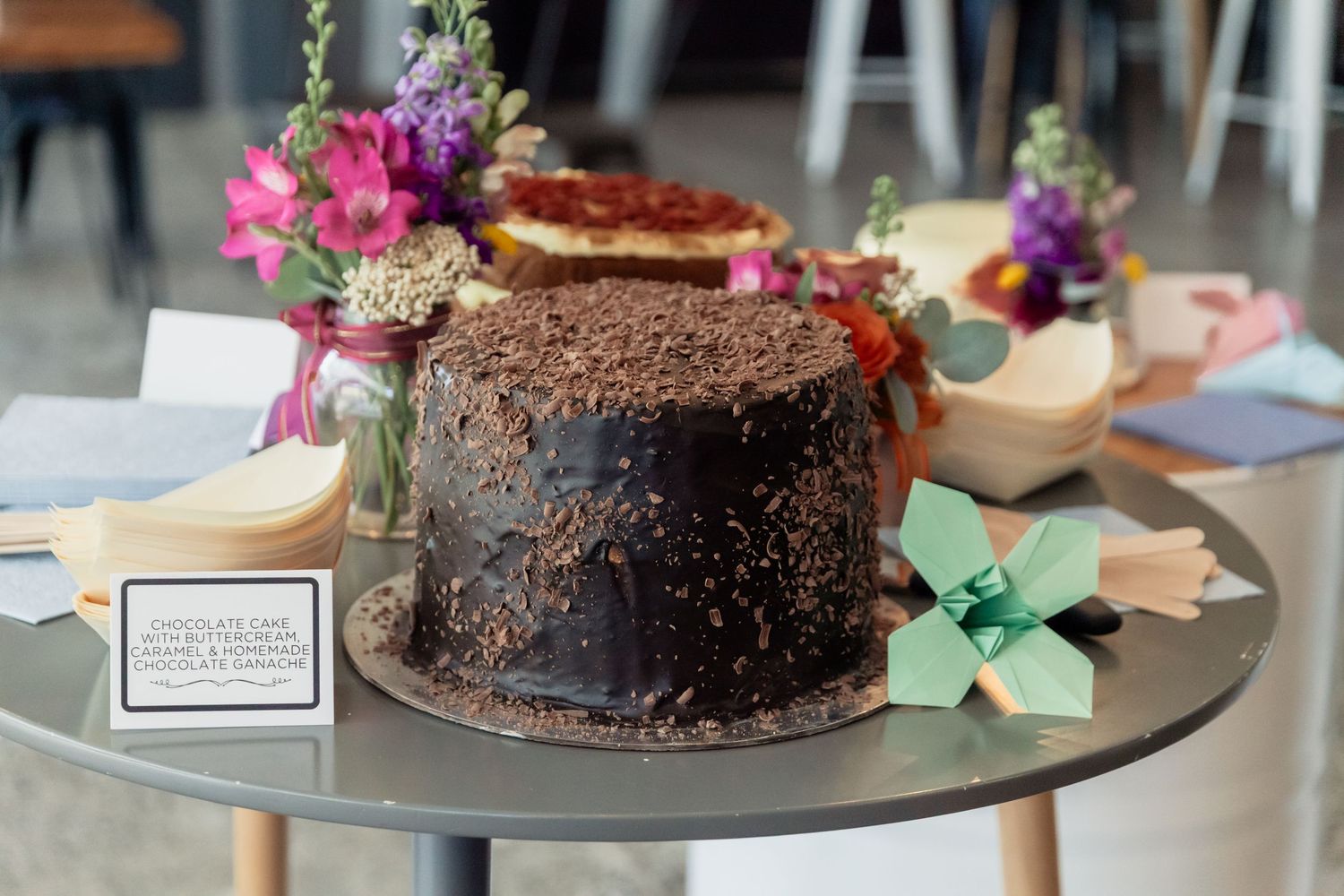 A rich chocolate cake with dark frosting sits on a silver serving plate surrounded by pink flowers and table decor.