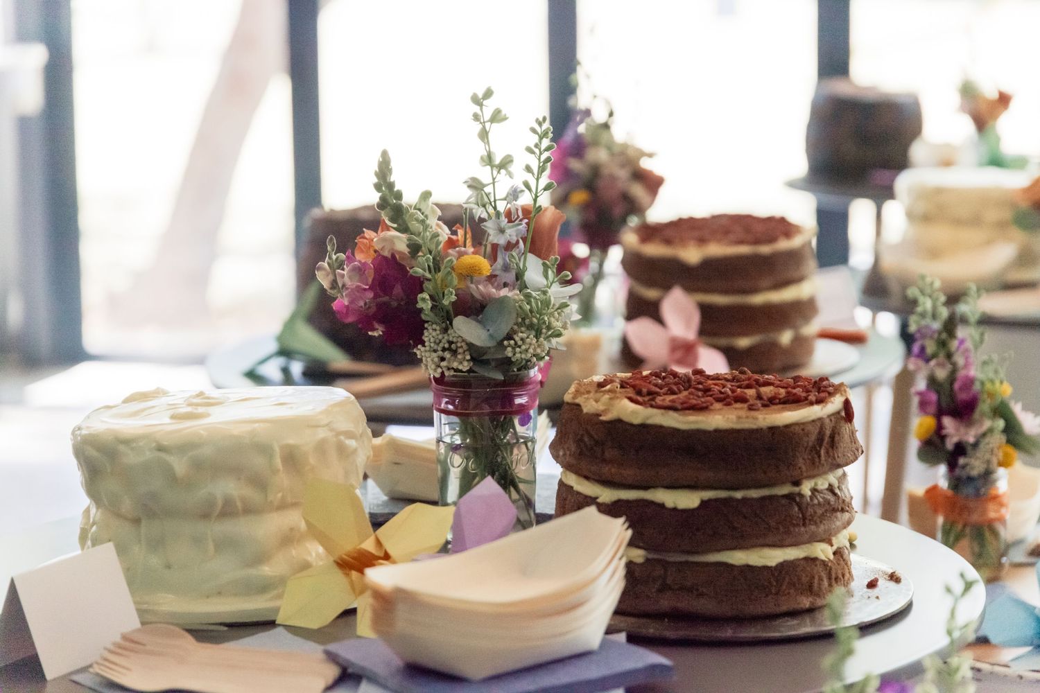 An assortment of decorated cakes and desserts displayed on a rustic wooden table with colorful floral arrangements.
