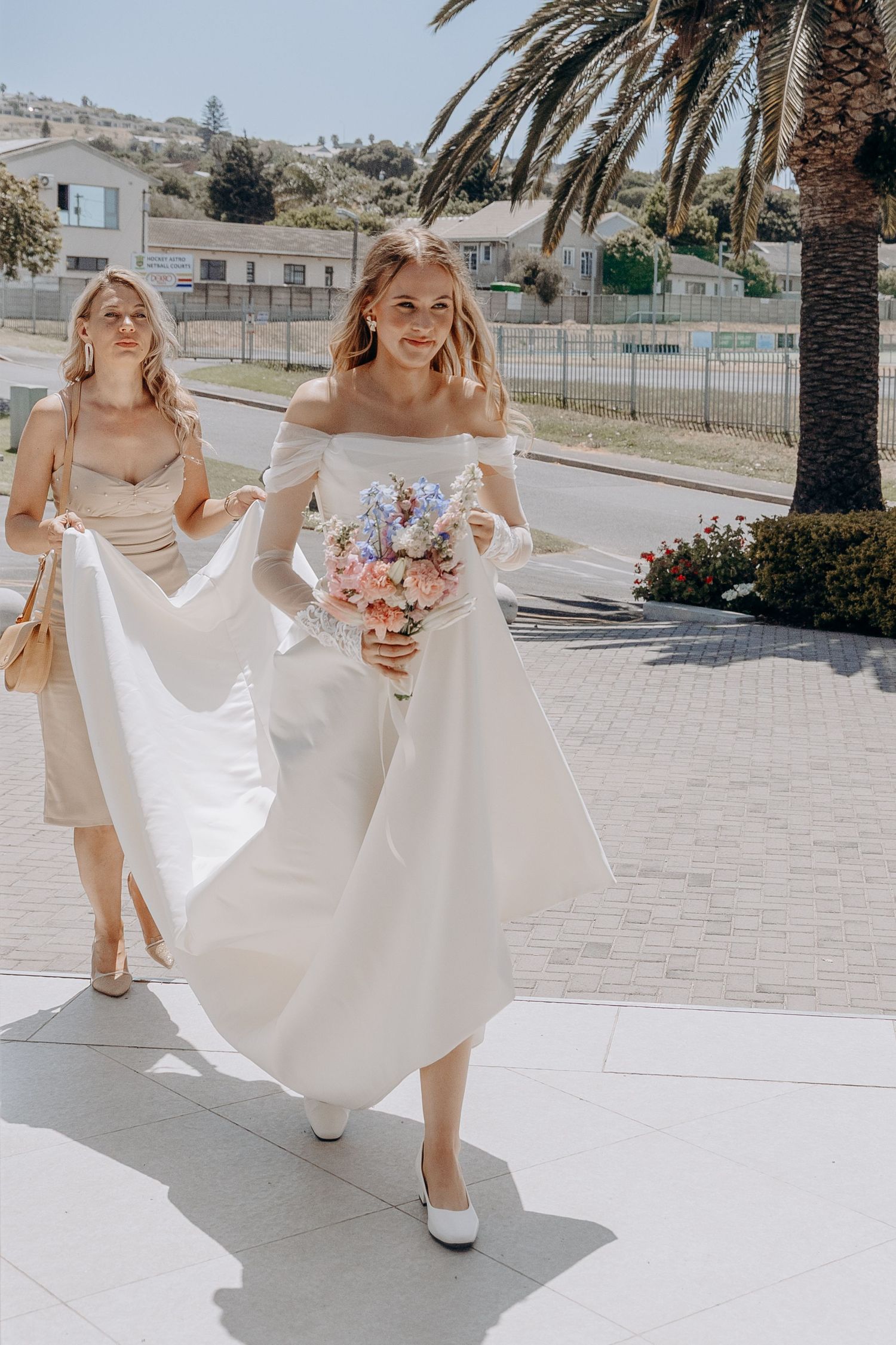 Wedding party members in white dresses walk together on a sunny paved path holding floral bouquets.