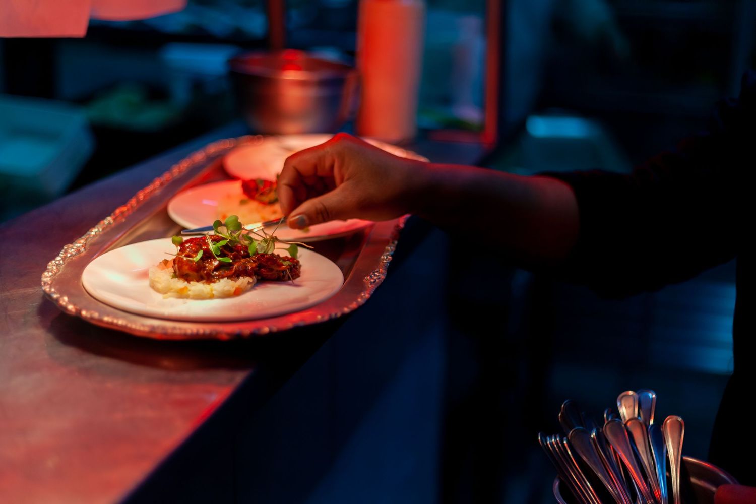 Hand placing garnished food on a white plate at a dimly lit restaurant counter with red ambient lighting.