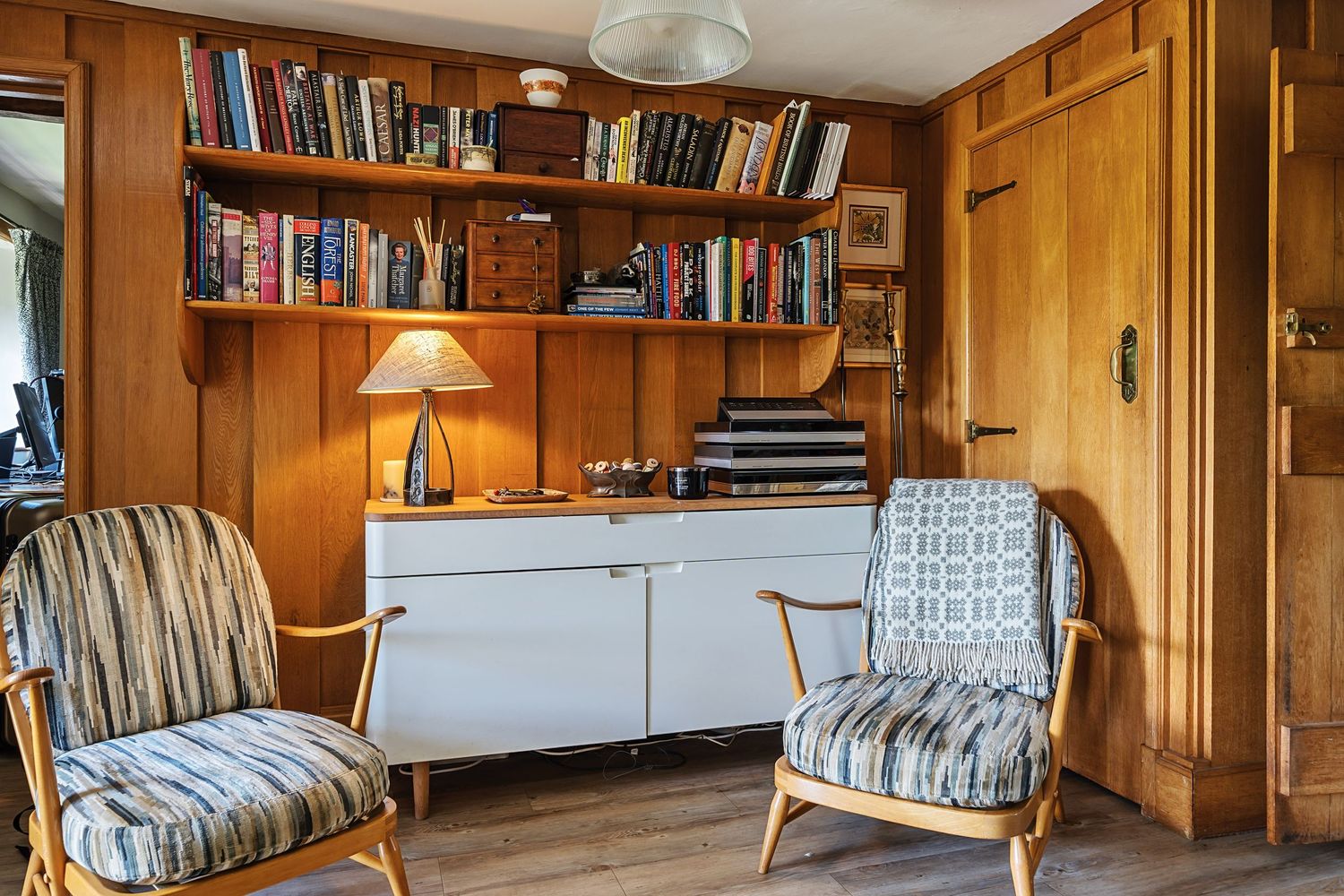 Cozy vintage reading nook with wood paneling, built-in bookshelves, retro chairs, and a white cabinet with table lamp.