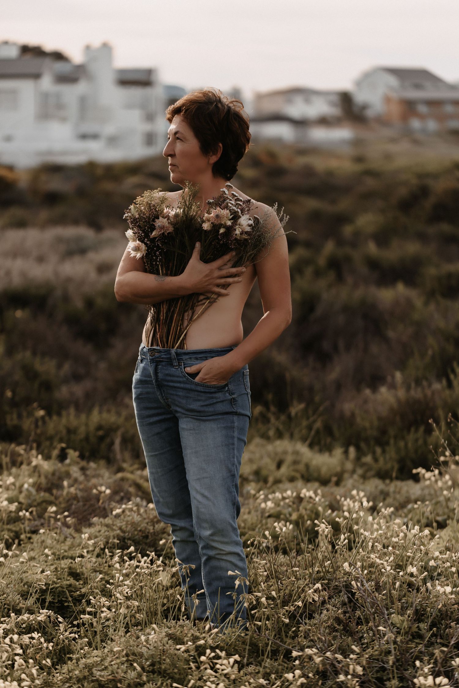 Two people embrace in a field of wildflowers at sunset with buildings visible in the background.