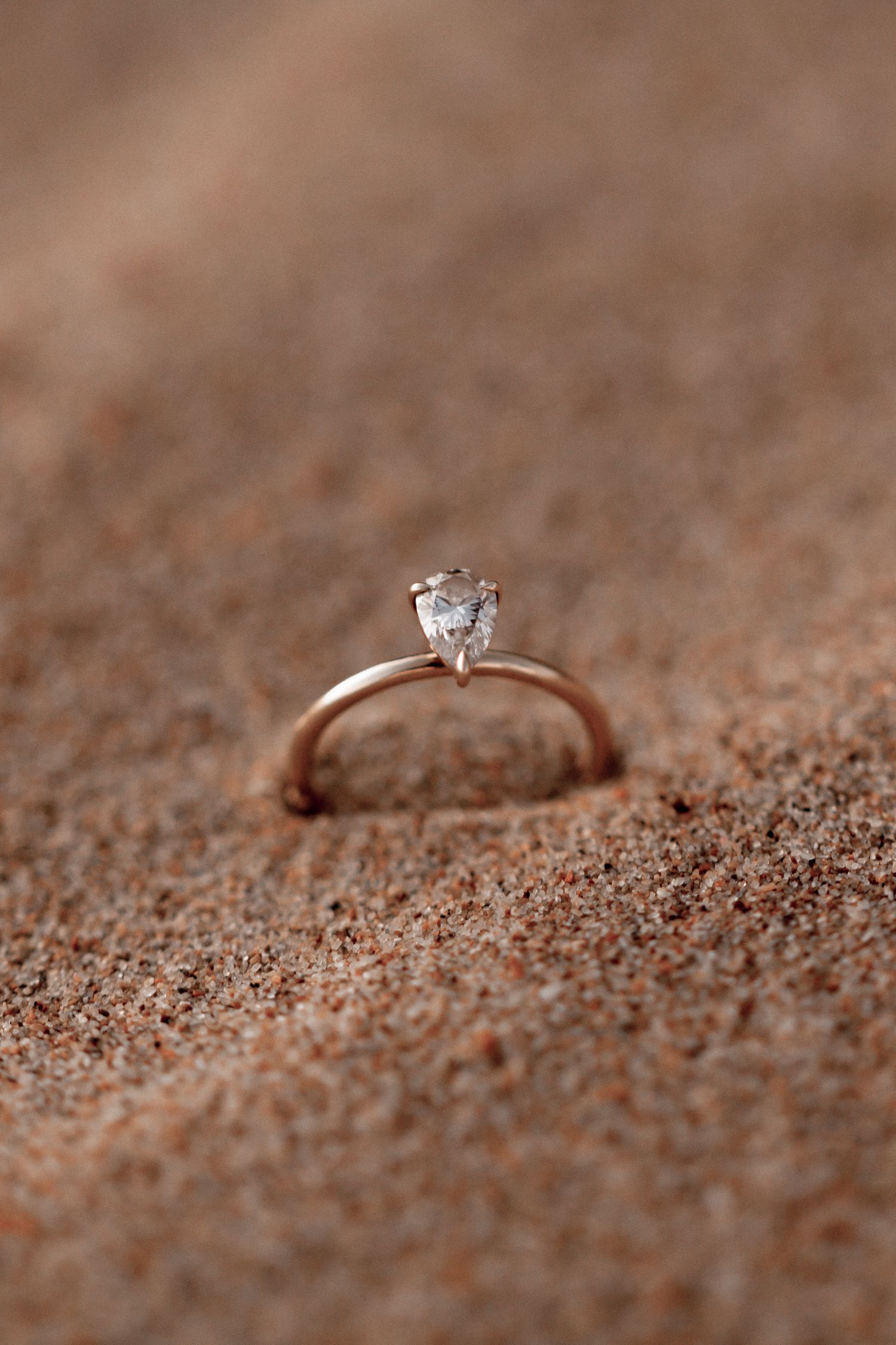 Diamond engagement ring sparkles against textured brown sand in a romantic beach proposal setting.