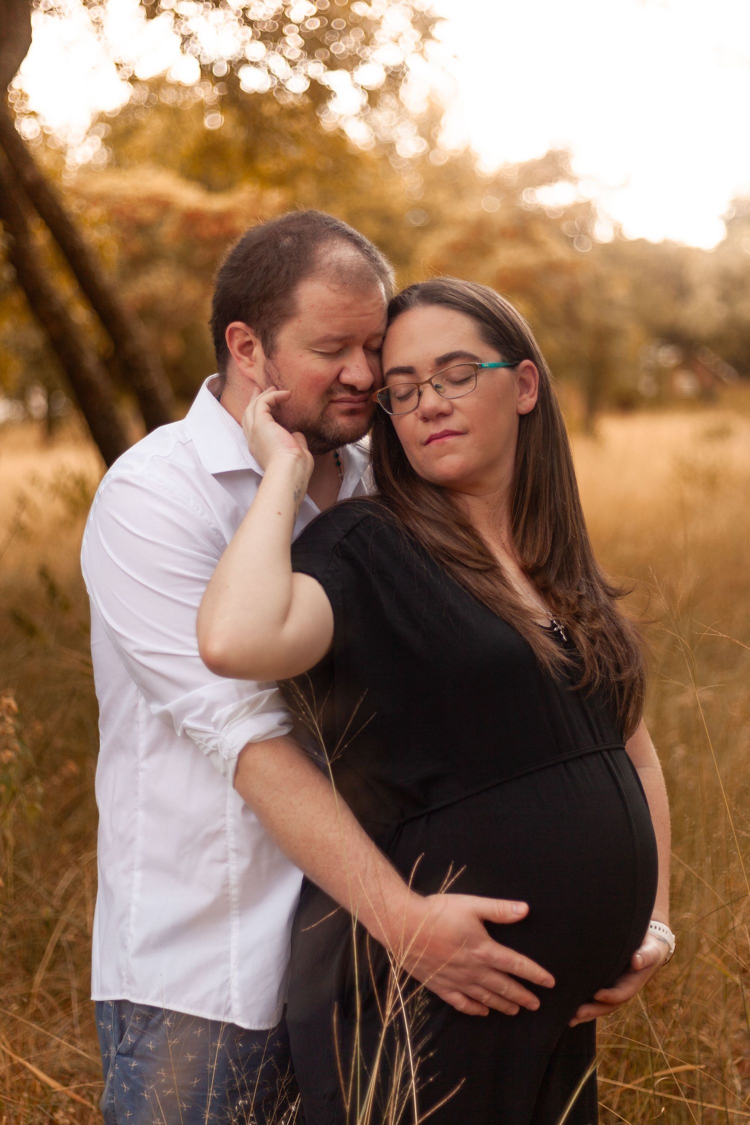 A couple embraces lovingly in a golden sunset field with tall grass during a romantic maternity photoshoot.