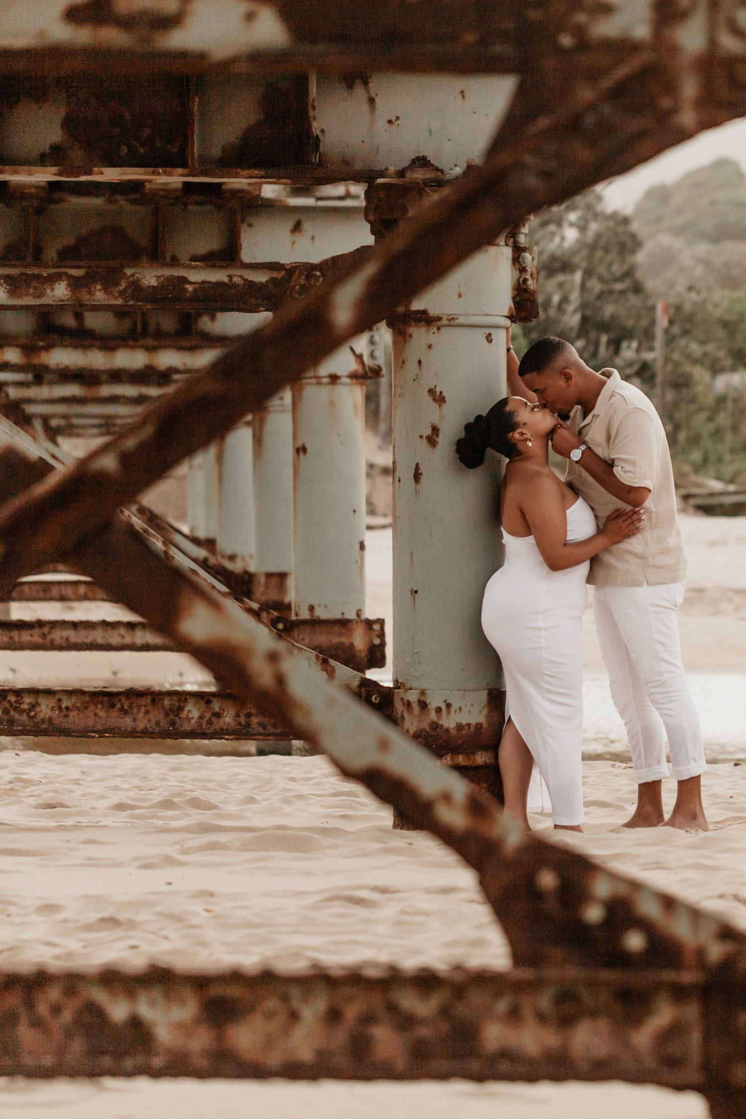 A couple in white outfits shares an intimate moment under a wooden pier at sunset.