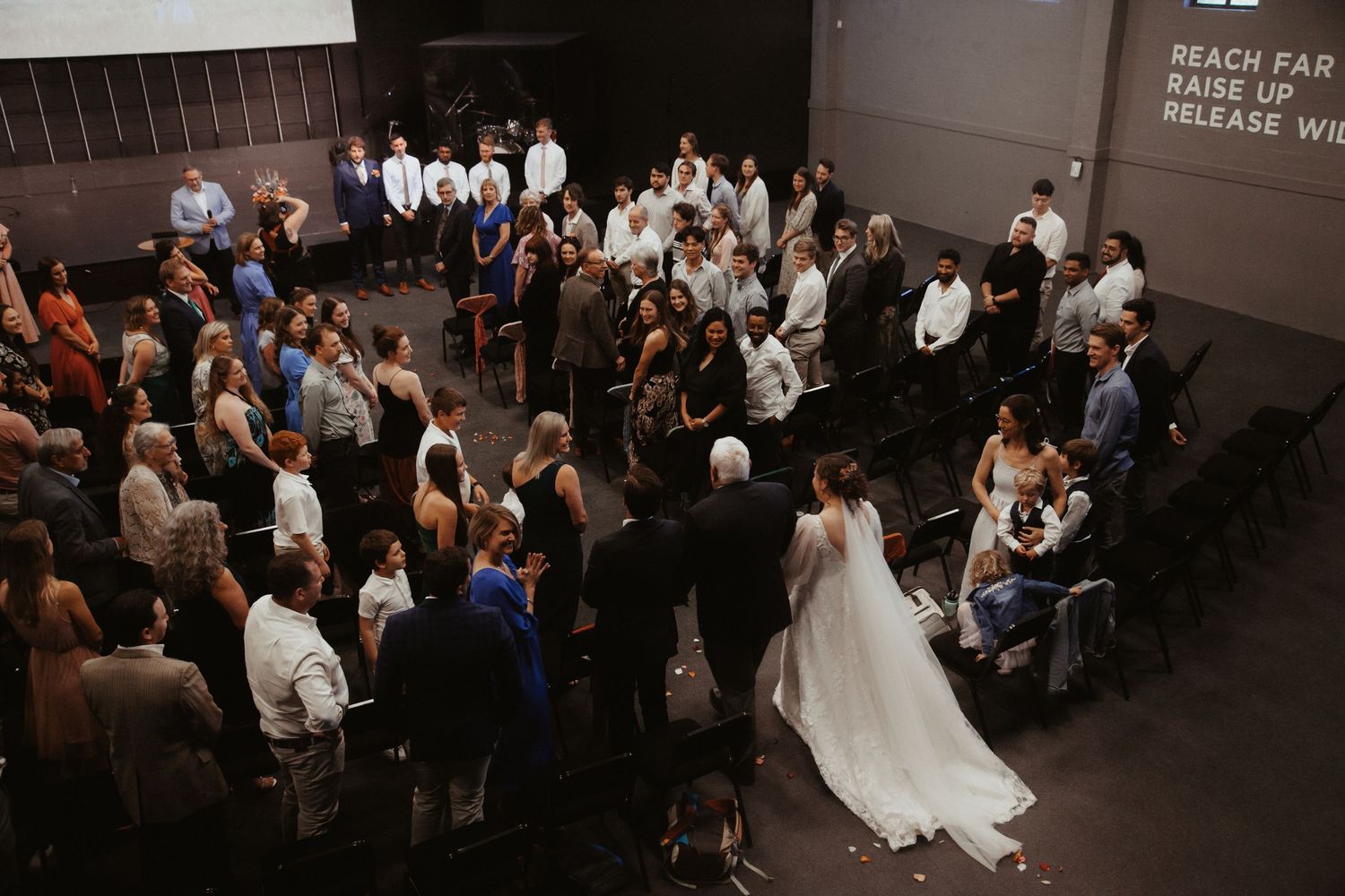 An overhead view of a wedding ceremony in a dark indoor venue with guests gathered around the couple in white.