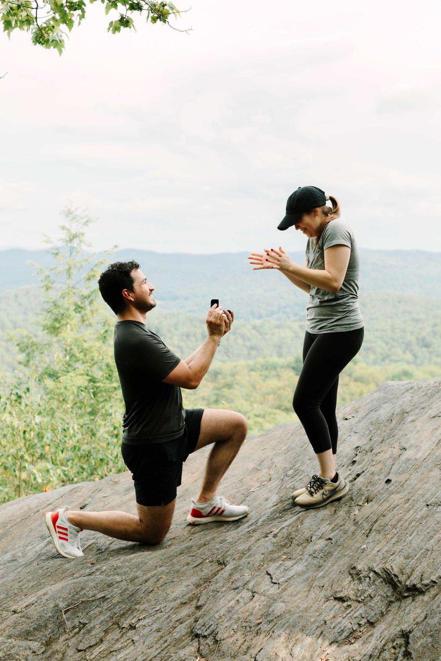 Man proposes on one knee to surprised woman on rocky overlook with lush green mountain valley behind them.