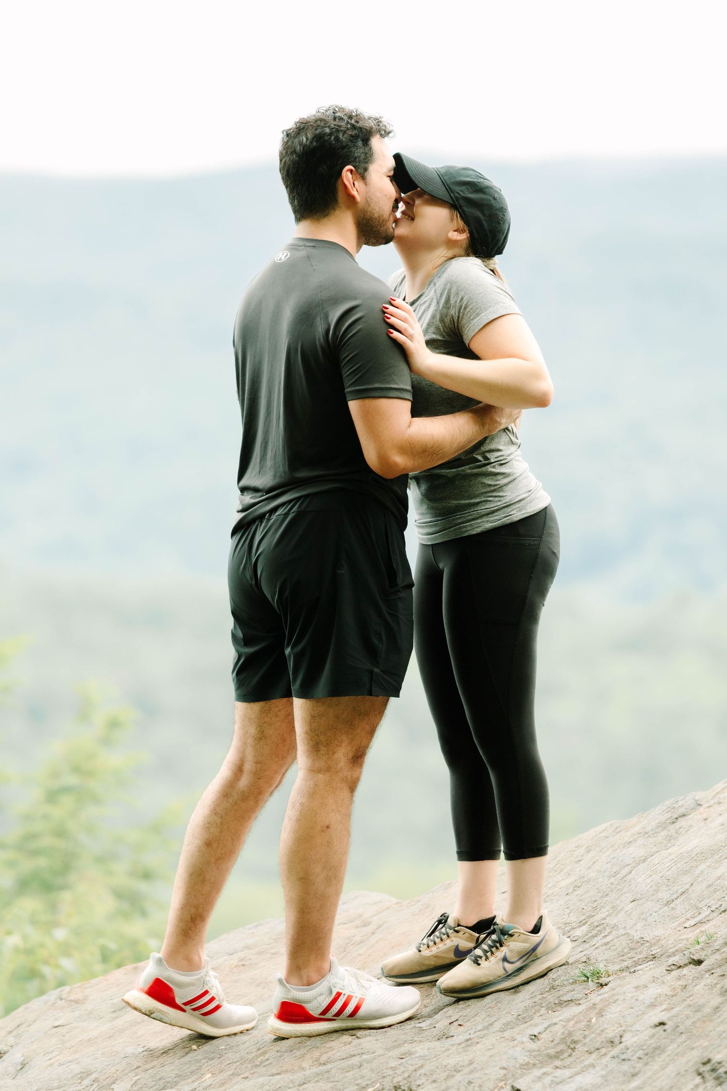 A couple sharing a romantic kiss on a rocky overlook with scenic mountain views in the background.