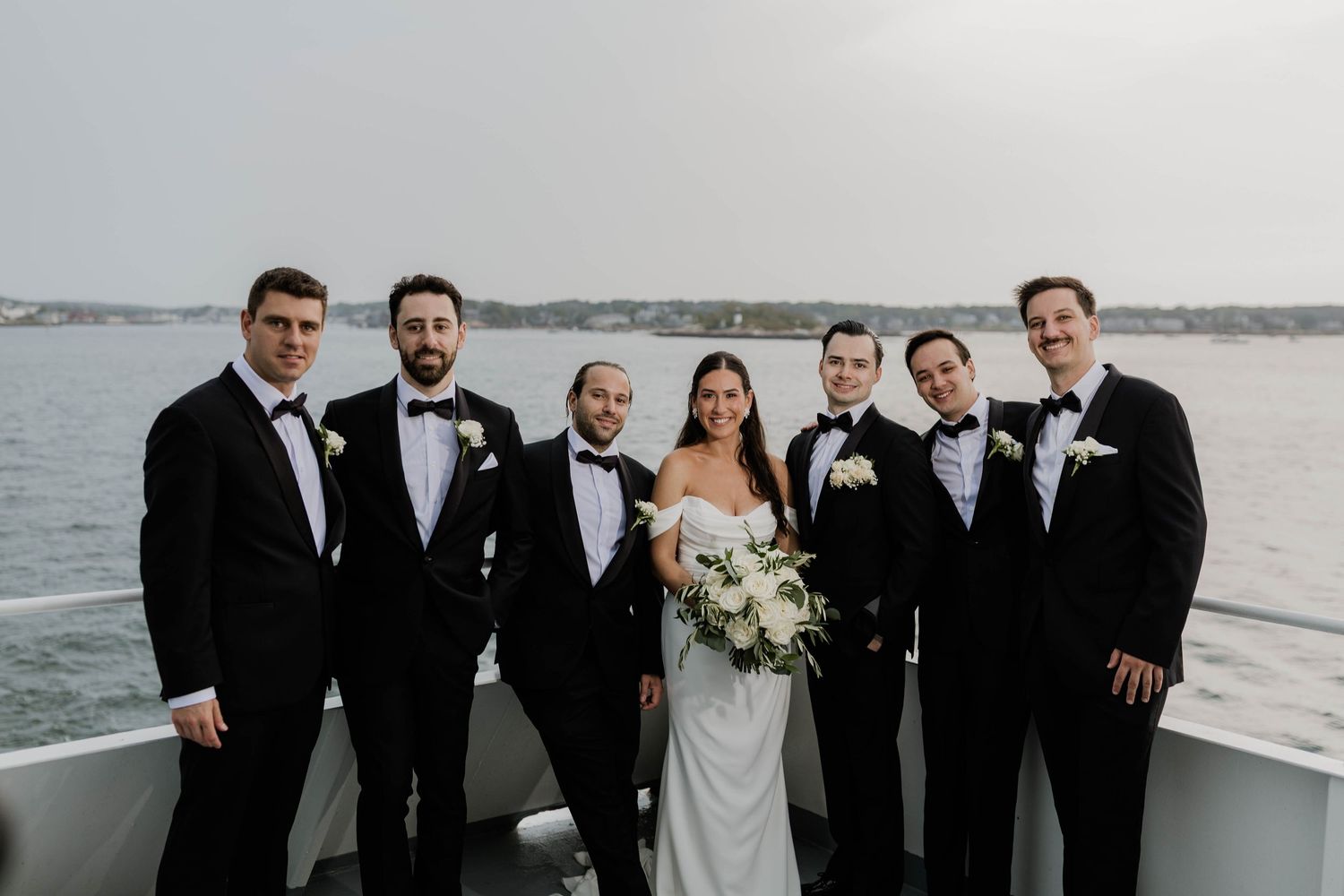 Bride and groom posing with groomsmen in black tuxedos on boat deck with water backdrop.